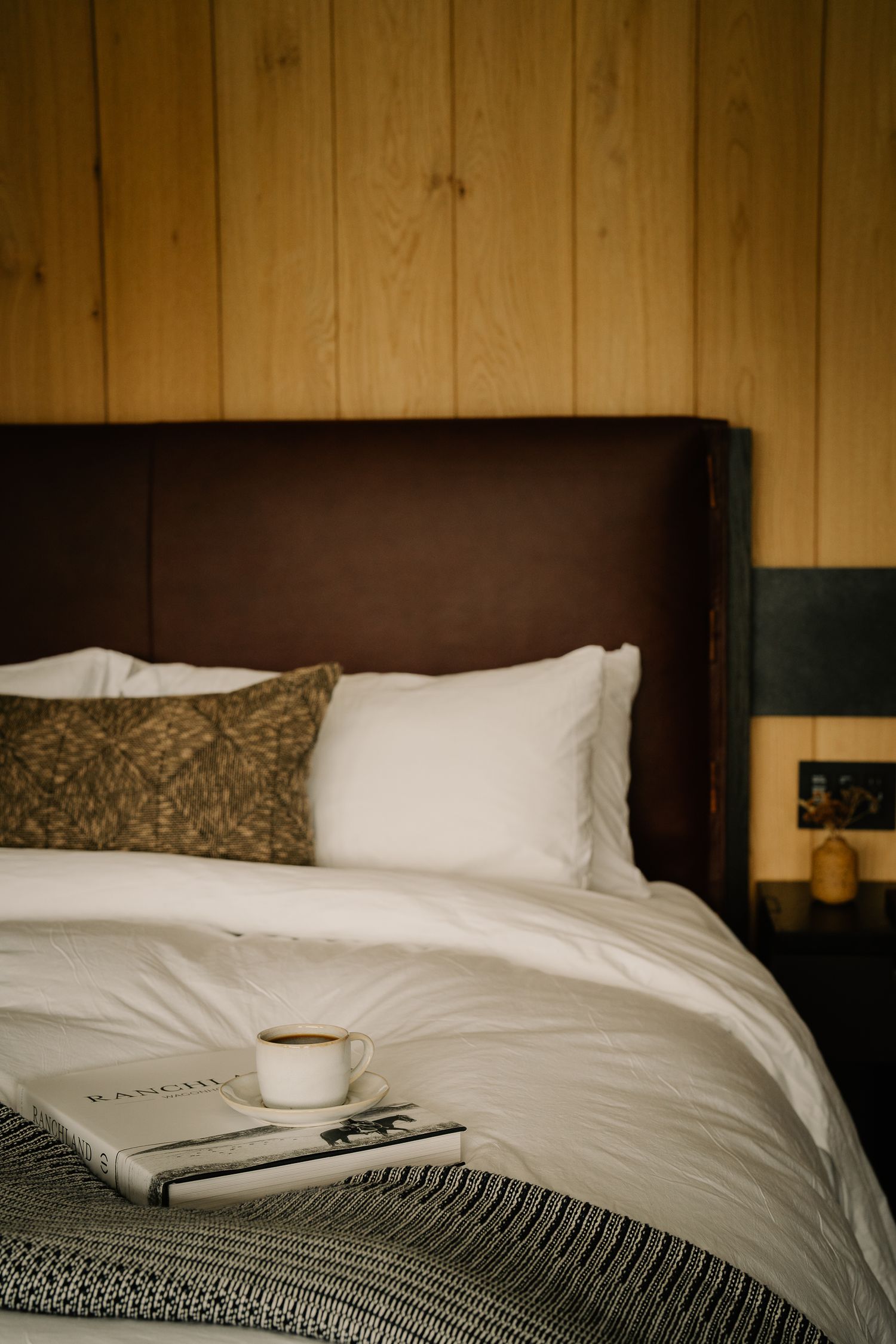 Cozy hotel bed with white linens, decorative pillows and coffee cup on magazine against wood paneled wall.
