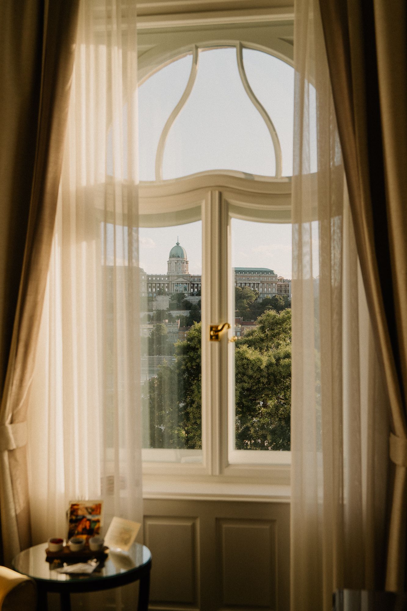Elegant arched window with sheer curtains overlooking Buda Castle and lush green trees in Budapest, Hungary.