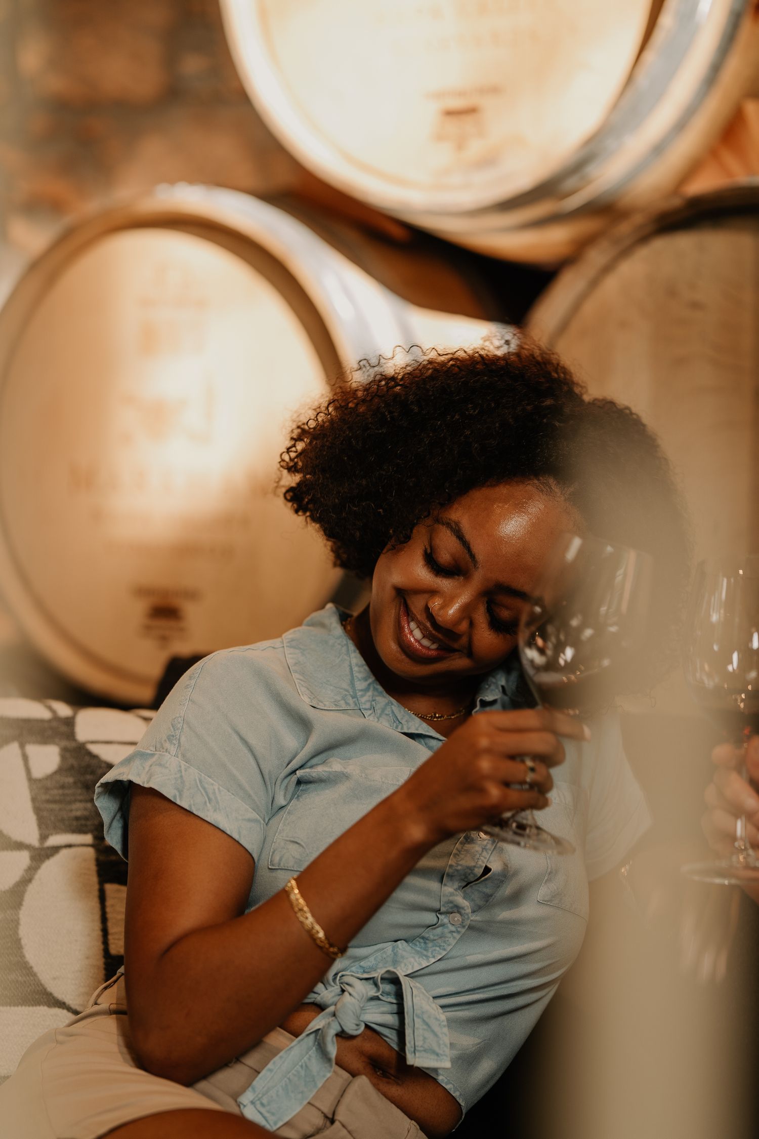A smiling woman relaxes among wine barrels in a cellar, holding a glass of red wine, wearing a denim dress and gold bracelet.
