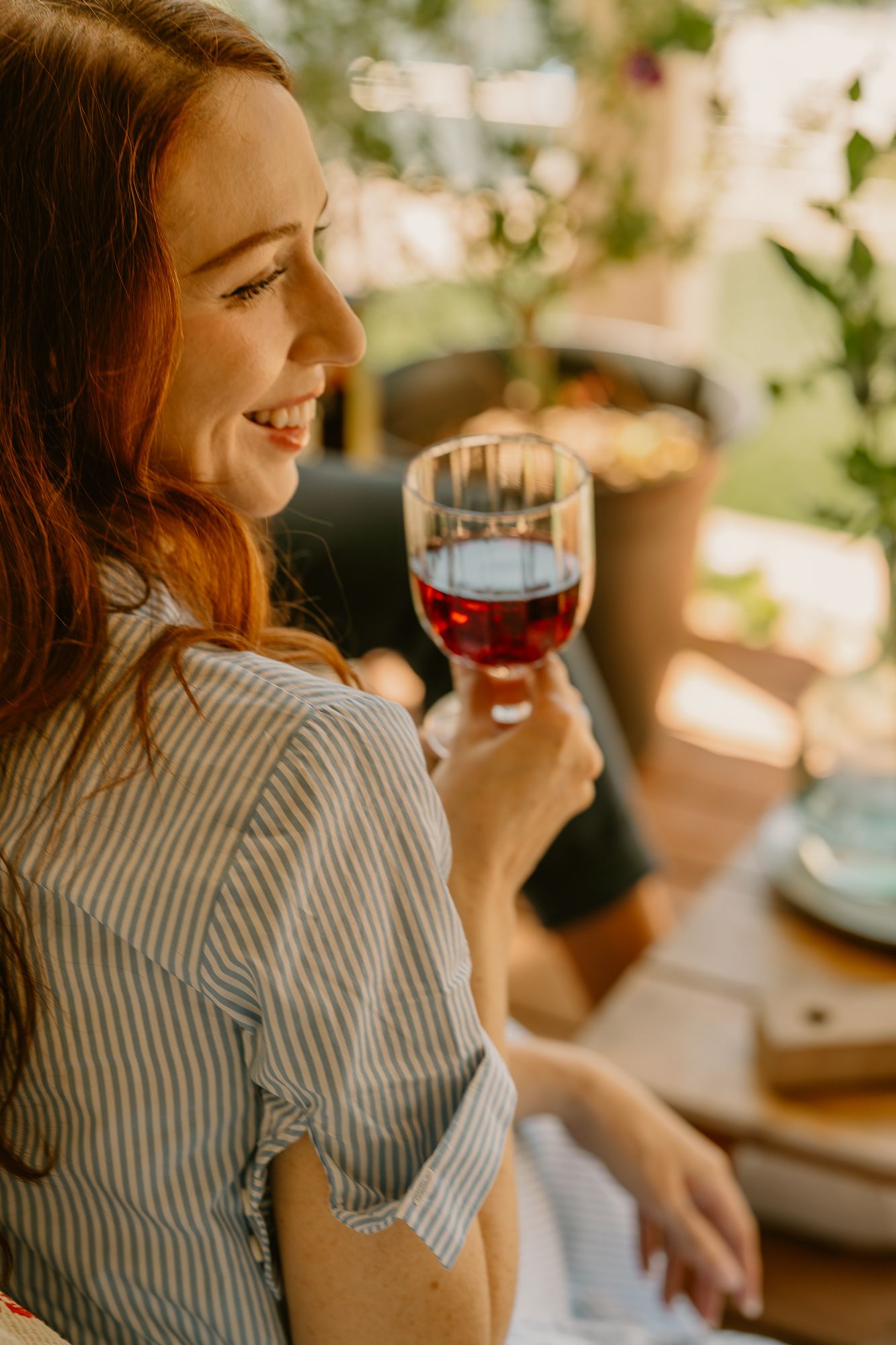 Smiling redhead woman enjoying a glass of red wine outdoors in warm golden sunlight.