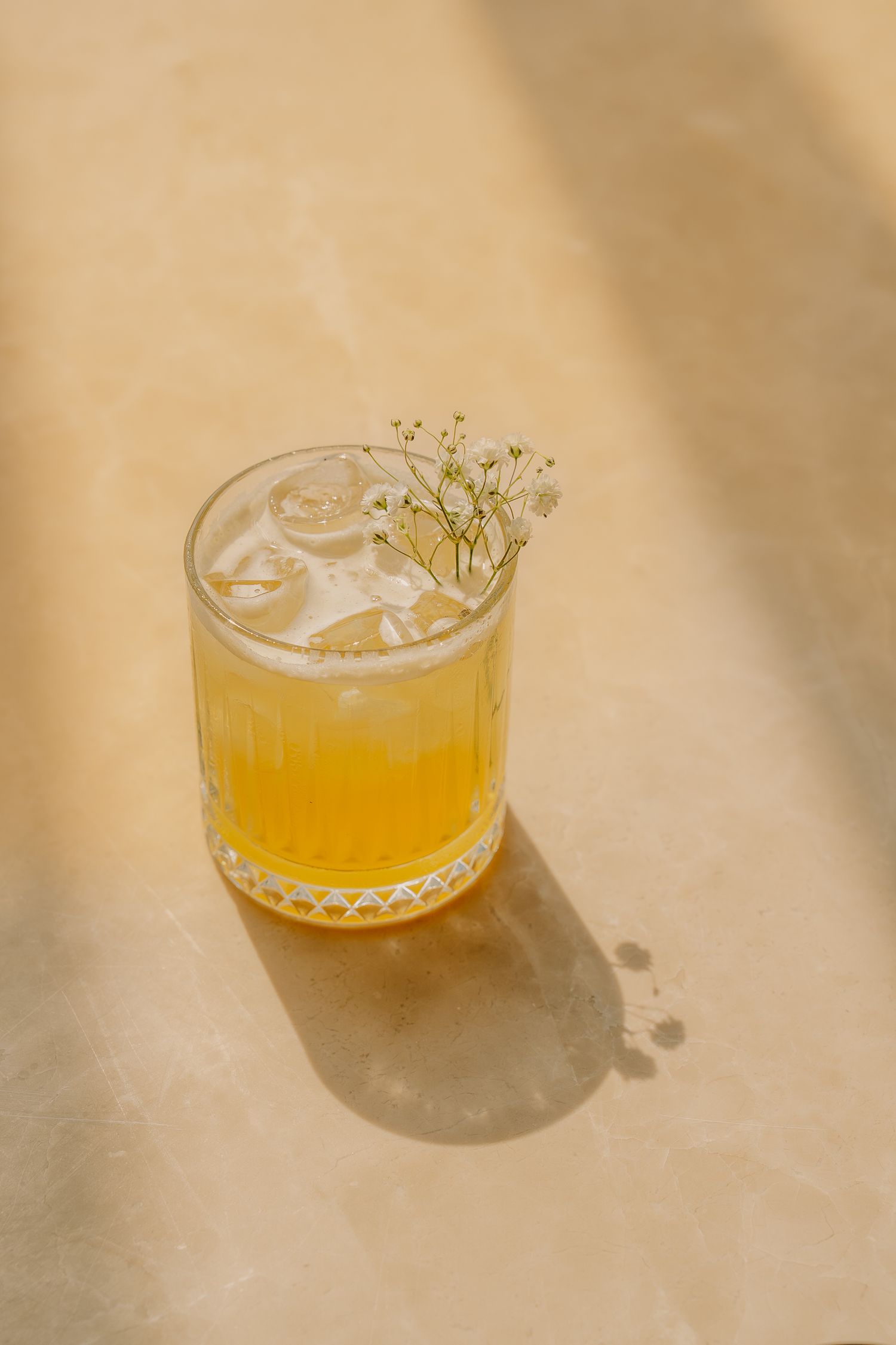 Golden cocktail with ice and baby's breath flower garnish in ribbed glass on warm beige surface.