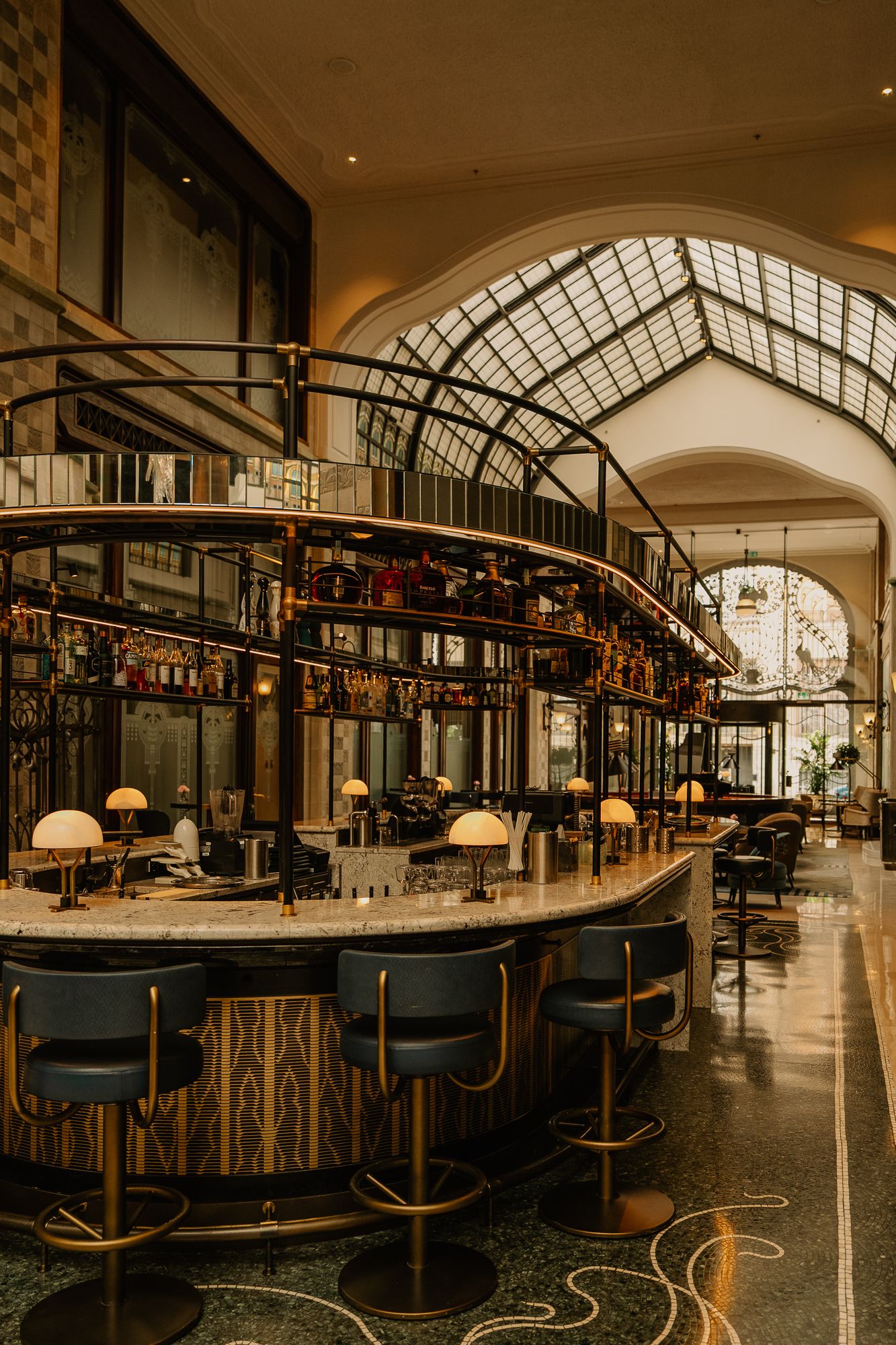 Elegant Art Deco bar with curved counter, gold accents, blue stools, and arched glass ceiling in a luxurious hotel lobby.