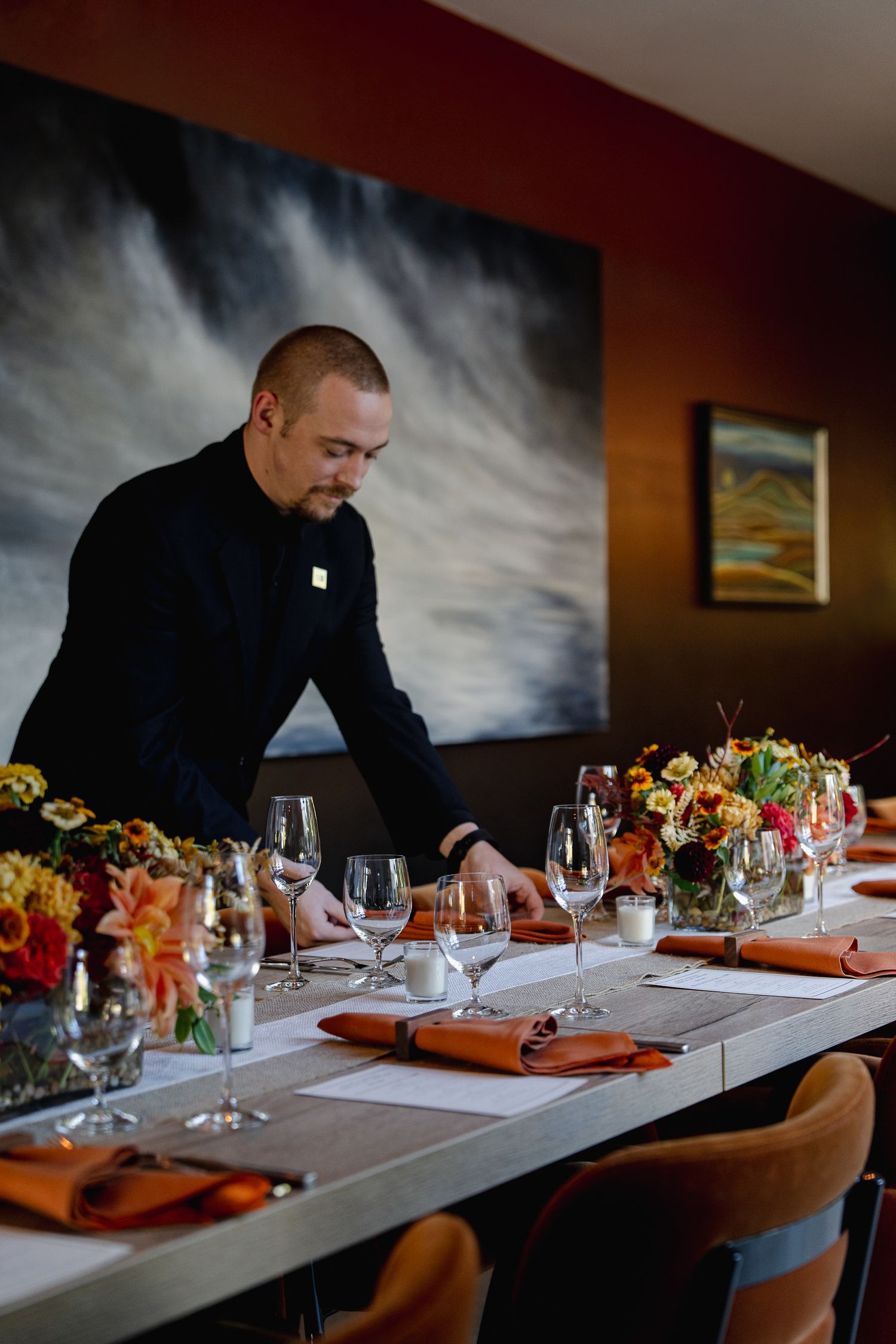 Restaurant staff member setting an elegant table with orange napkins and floral centerpieces against a dramatic wall mural.