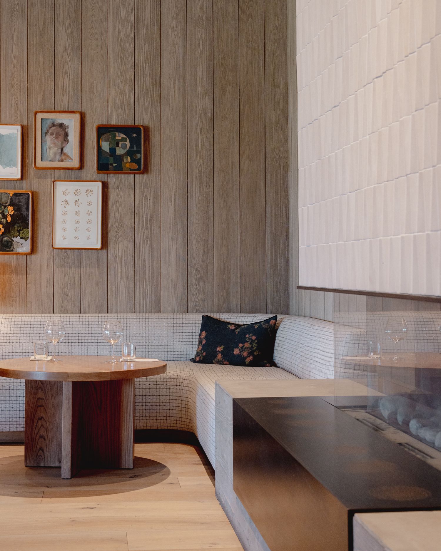 Modern restaurant interior with built-in corner bench seating, round wooden table and framed artwork on wood-paneled walls.