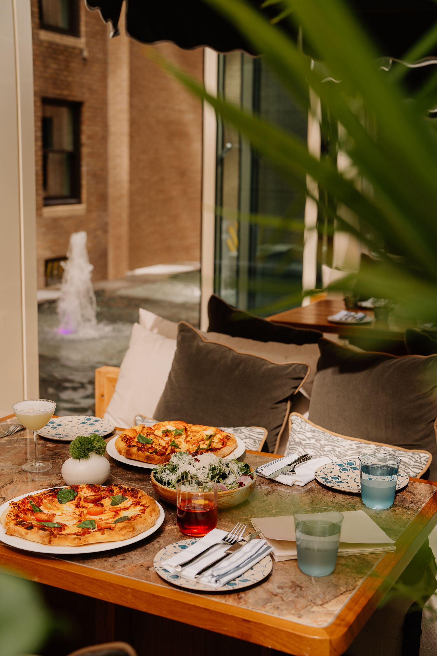 Cozy outdoor dining setting with fresh pizzas, wine glasses, and comfy cushions framed by plants and a fountain view.