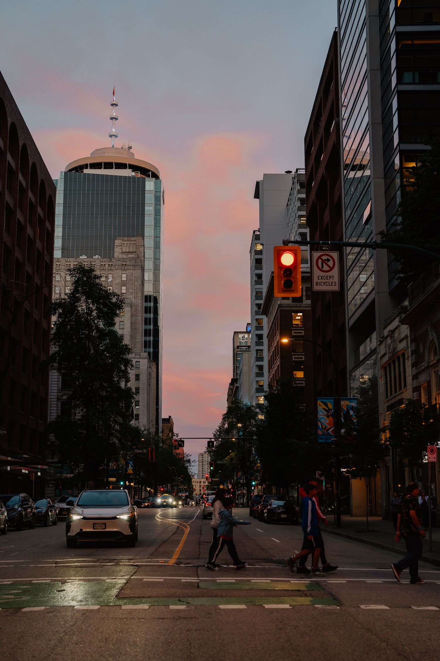 Urban street scene in downtown Vancouver at sunset with One Bentall tower and pedestrians crossing against pink sky.