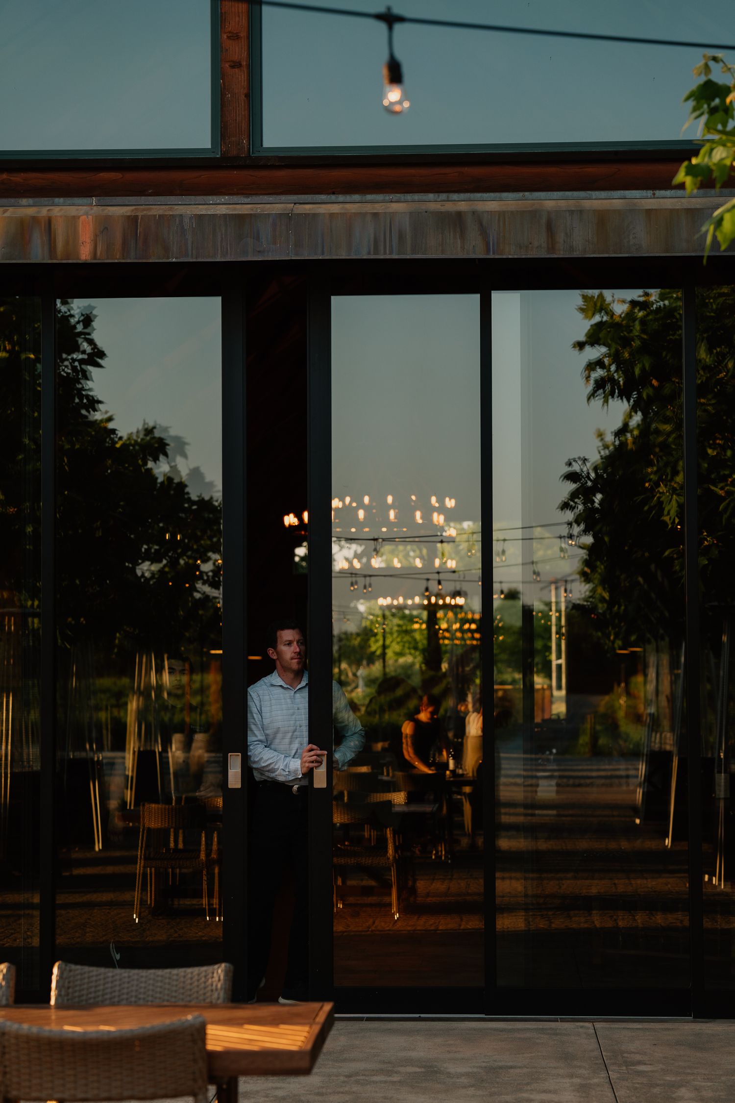 Evening view through glass windows of a modern restaurant with warm lighting and wooden furnishings.