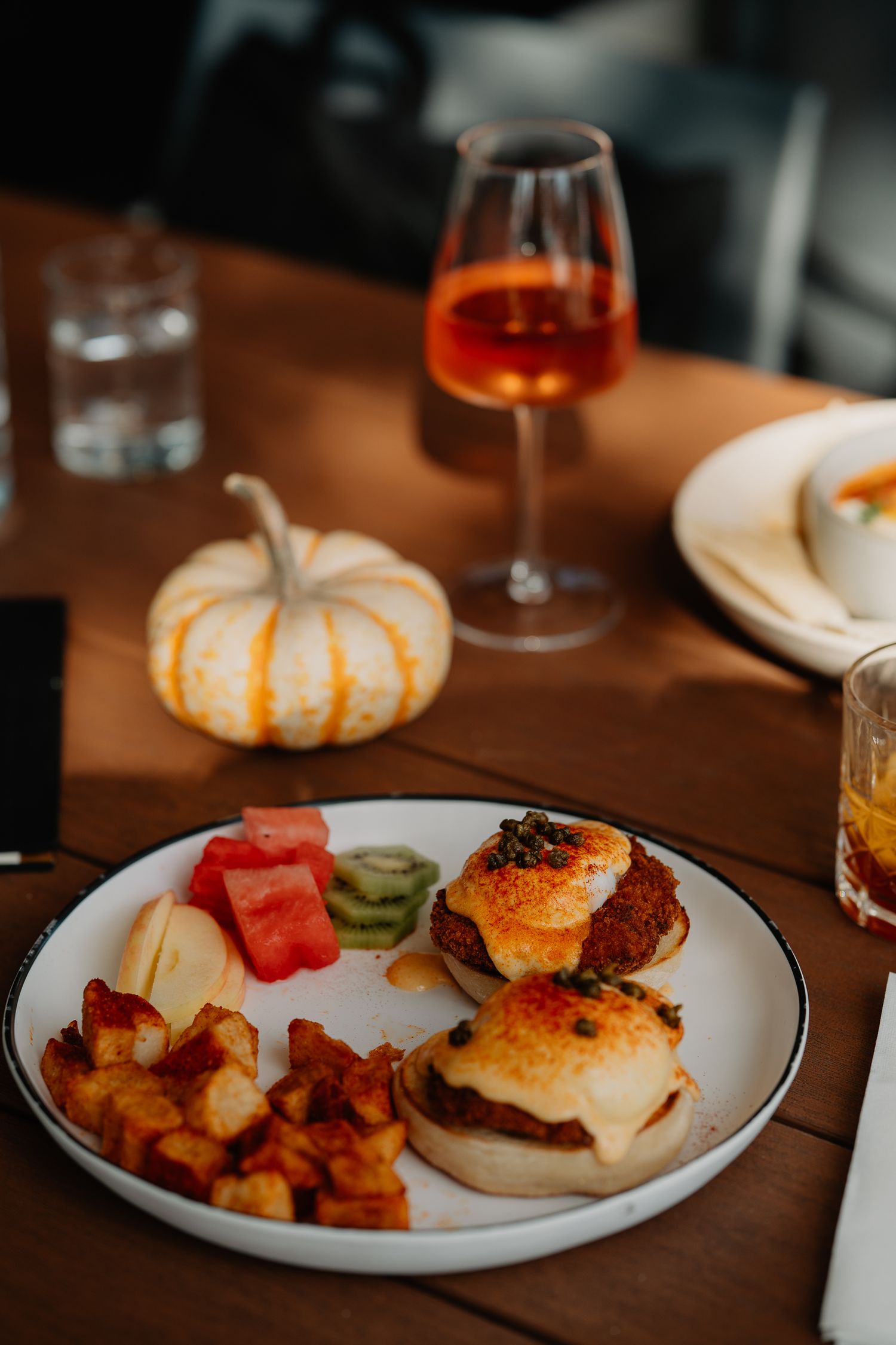 A cozy fall brunch setting with sliders, roasted potatoes, fruit, and a glass of rosé wine next to a decorative pumpkin.