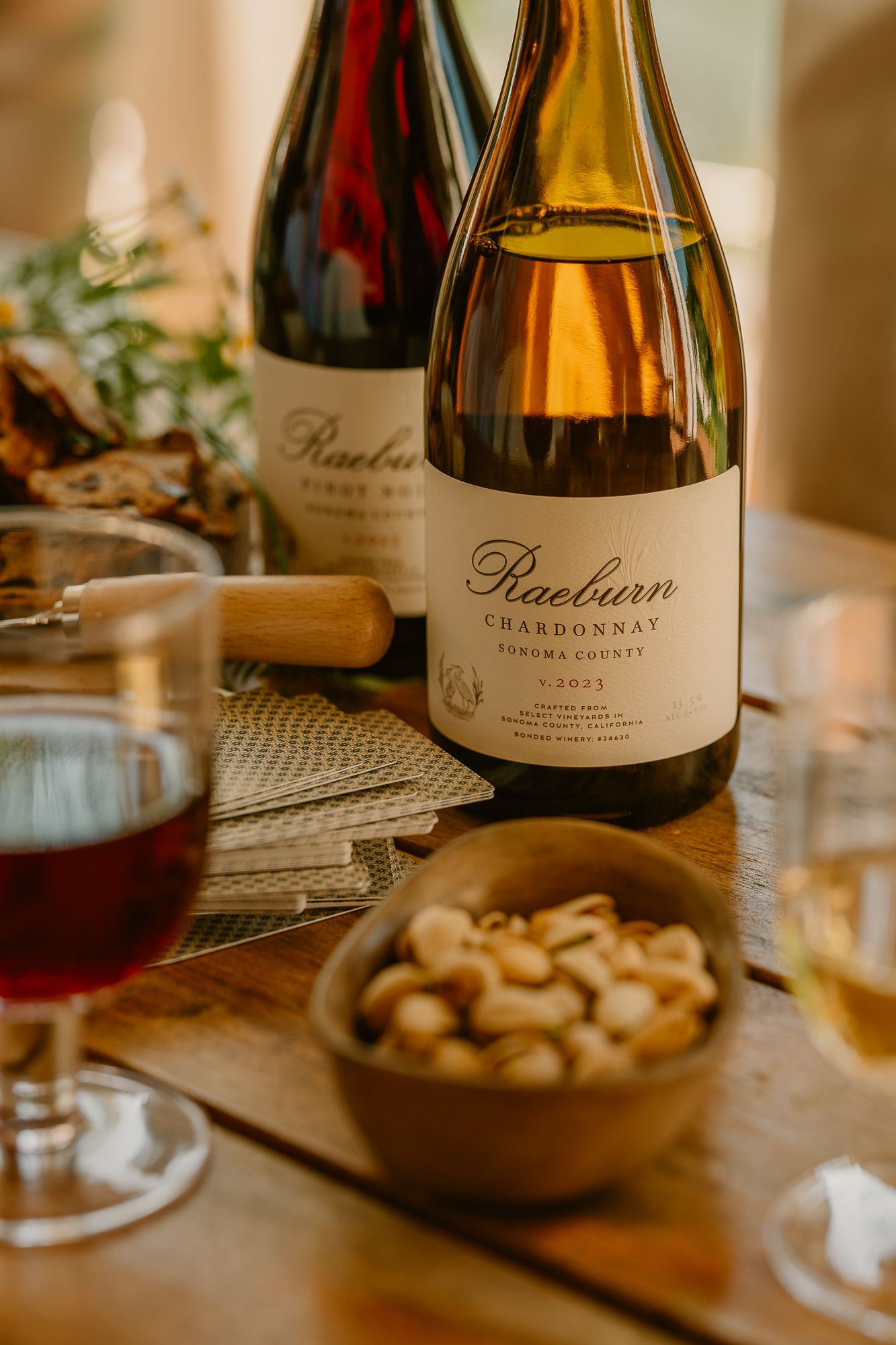 Raeburn Chardonnay and Pinot Noir wine bottles on a wooden table with cashews and a glass of red wine.