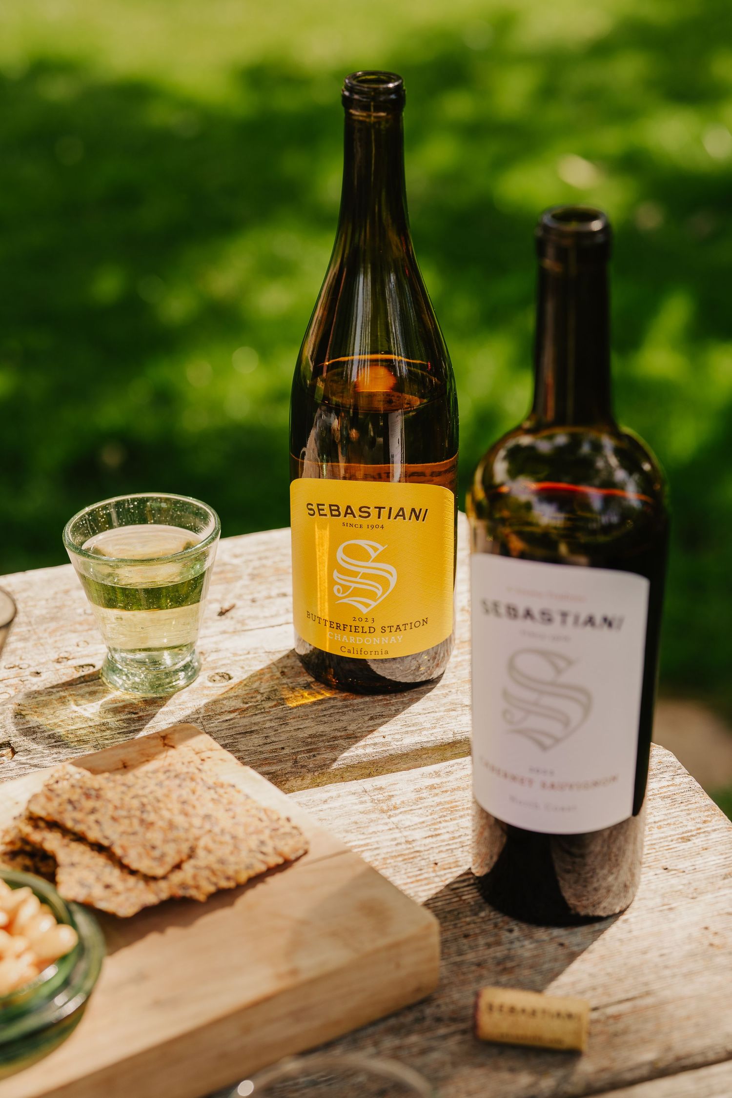 Two Sebastiani wine bottles on a wooden table outdoors with crackers and a glass of white wine.