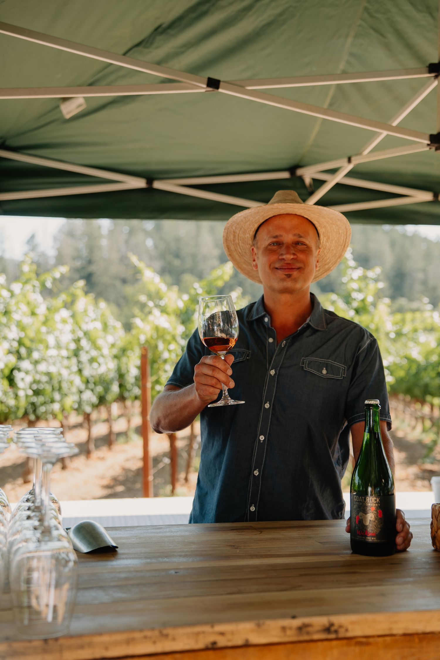 A wine tasting under a green canopy tent with vineyard rows visible in the sunny background.