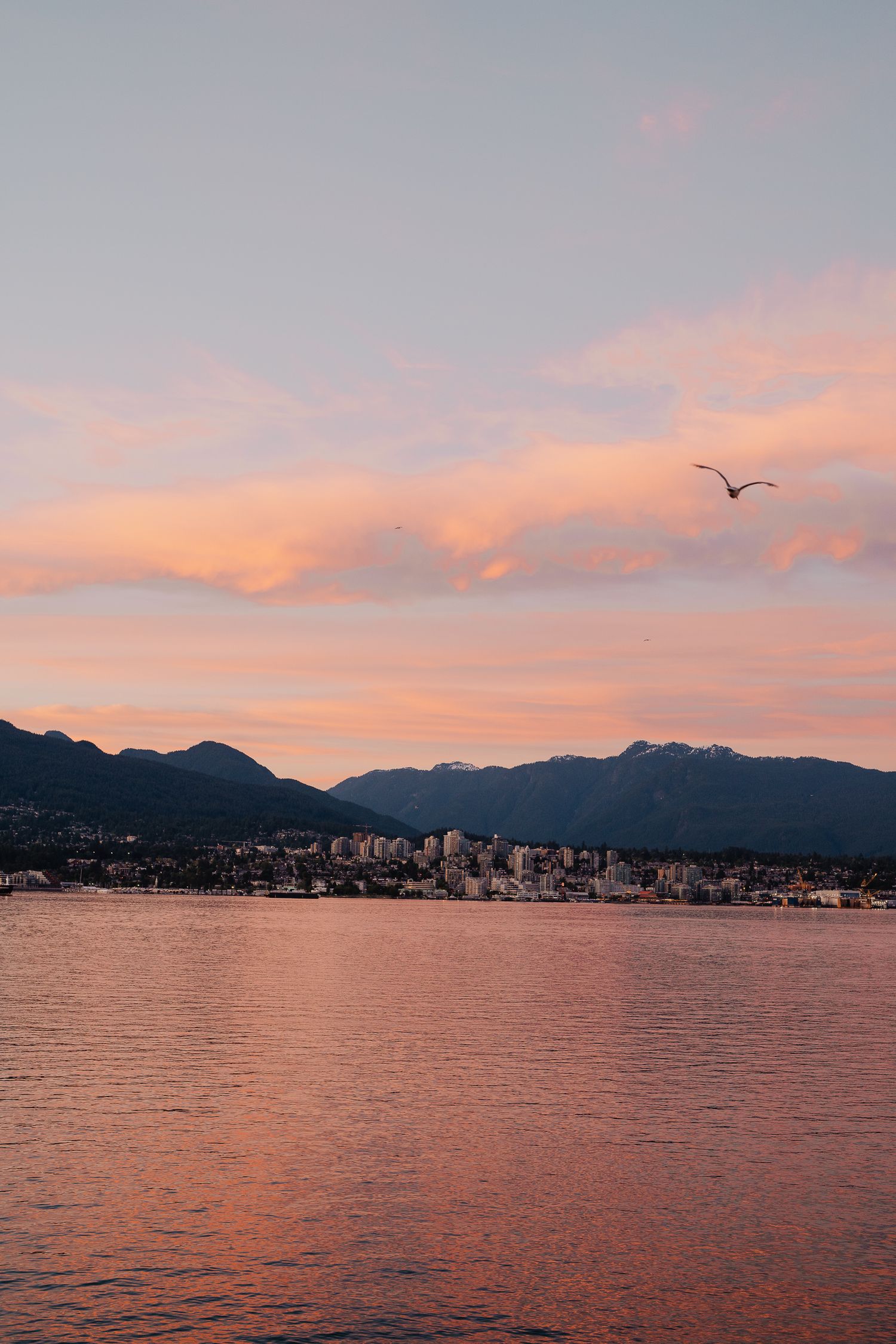 Sunset over Vancouver harbor with pink clouds reflecting on water and mountains silhouetted against orange sky.