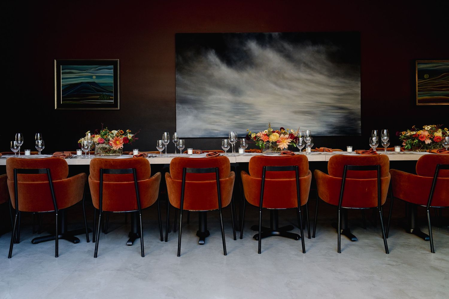 A long dining table with red velvet chairs set against a dramatic dark stormy landscape painting on the wall.