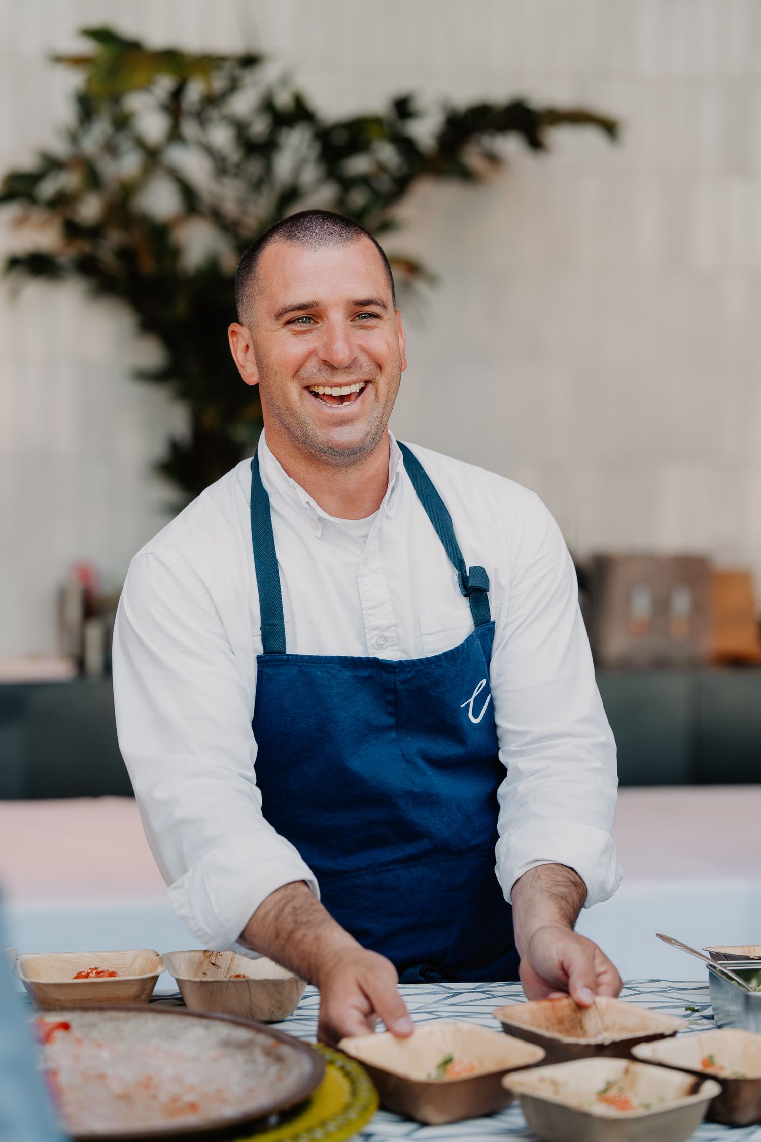 Smiling chef in white coat and blue apron prepares dishes at an outdoor catering station.
