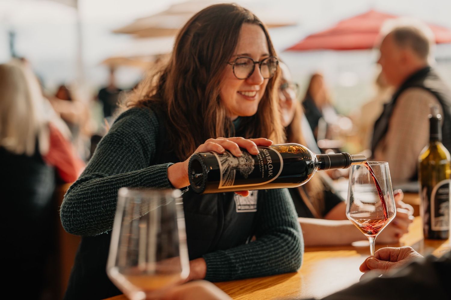 Smiling woman with glasses pouring red wine into a glass at an outdoor wine tasting event.