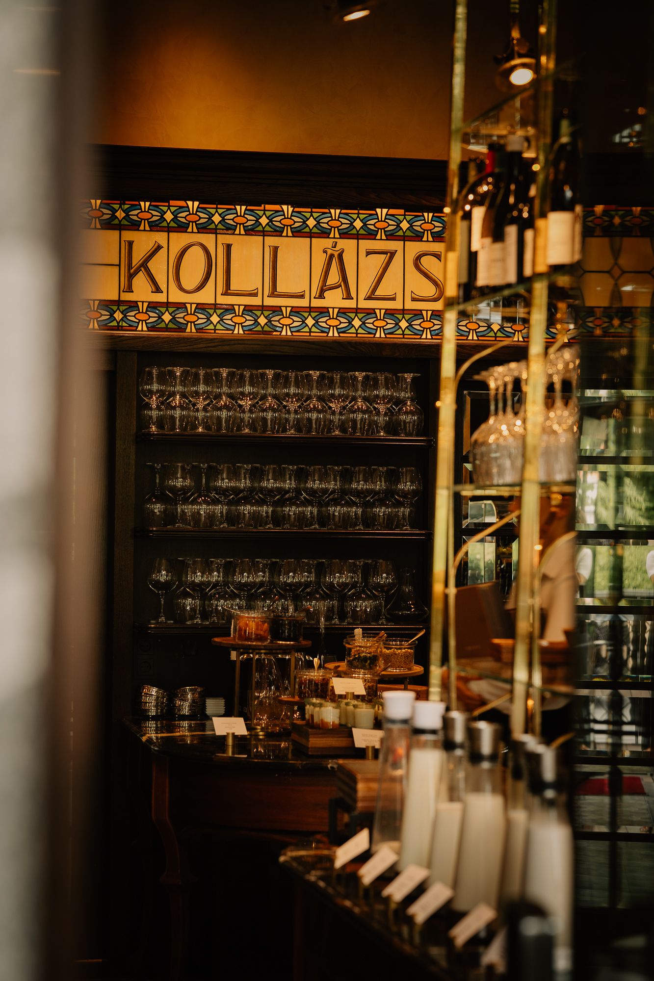 Interior view of Kollazs bar with elegant glassware display, golden accents, and warm ambient lighting.