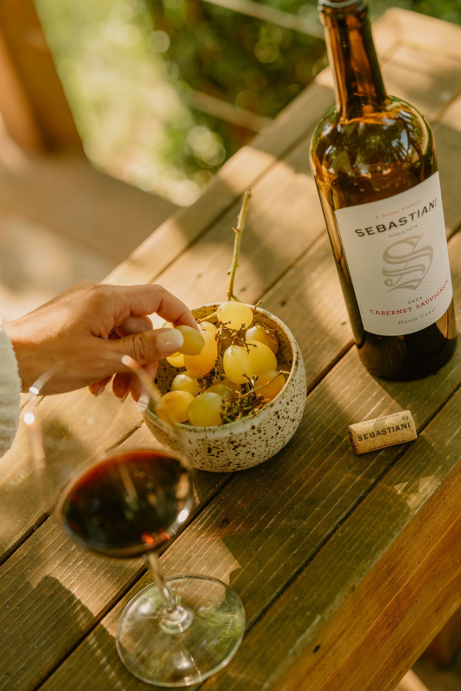 Hand reaching for fresh green grapes beside a glass of red wine and Sebastiani Cabernet Sauvignon bottle.