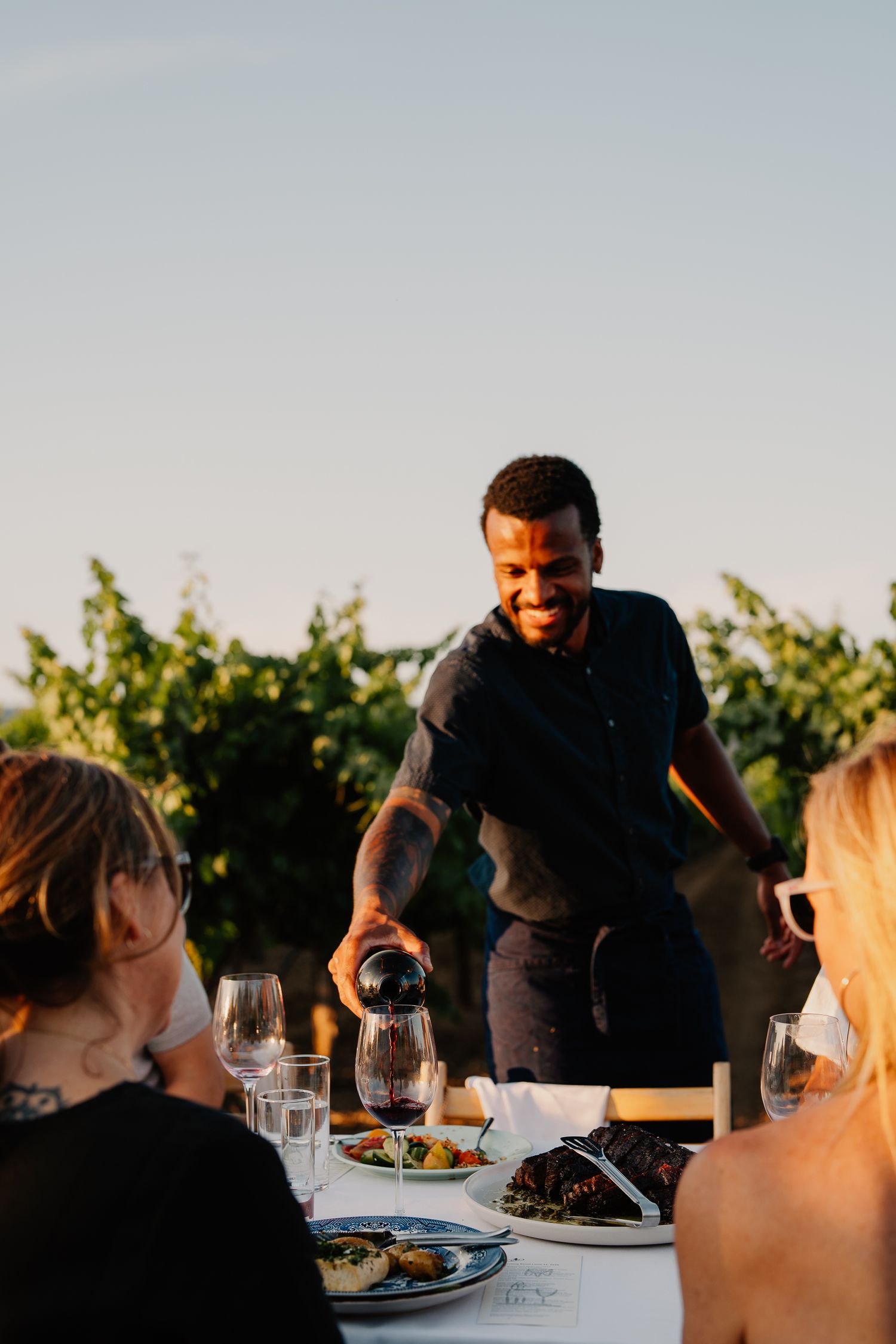 Server pours wine for guests at an outdoor vineyard dinner table during golden hour.
