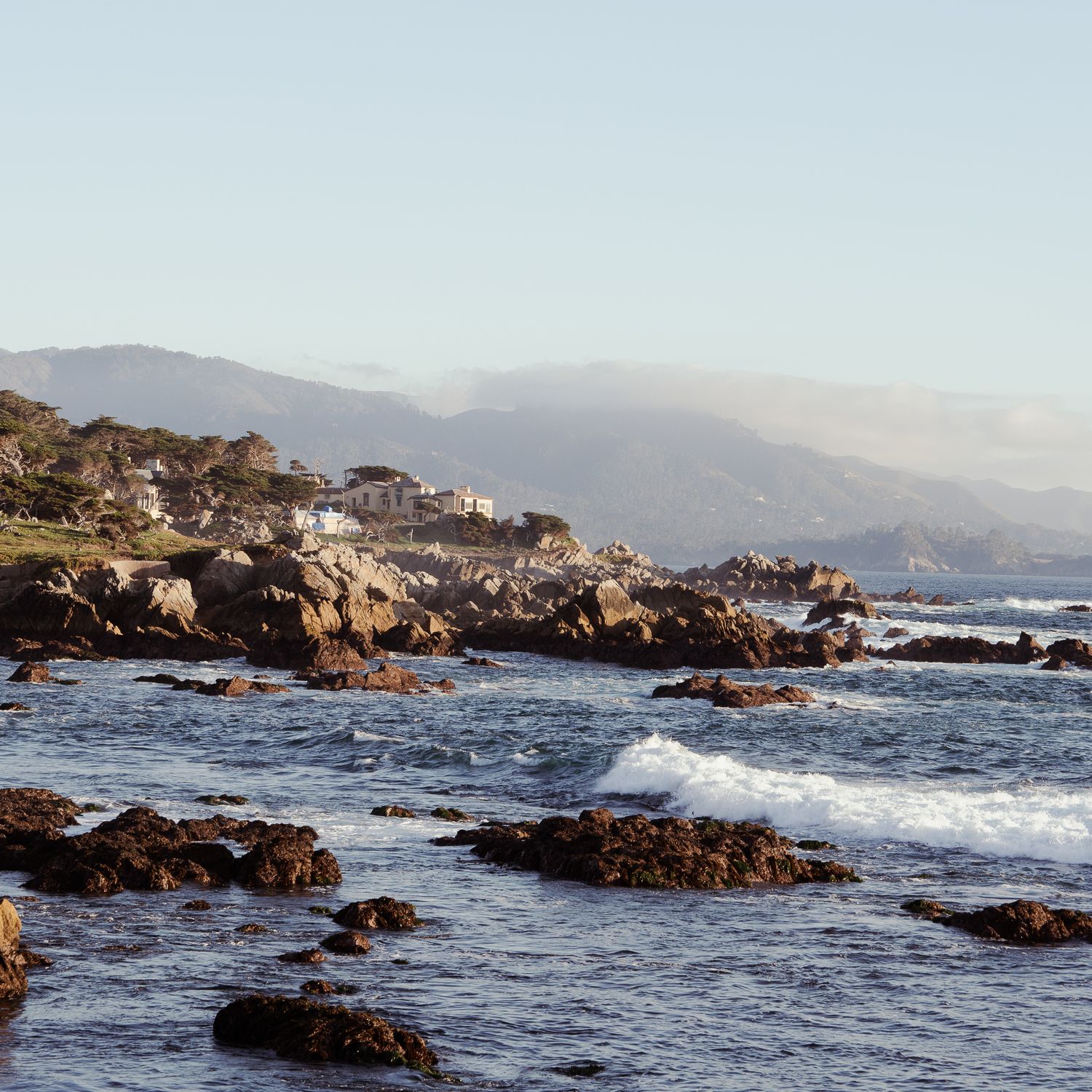 Rocky coastline with crashing waves against jagged outcrops along the Pacific Ocean during sunset.