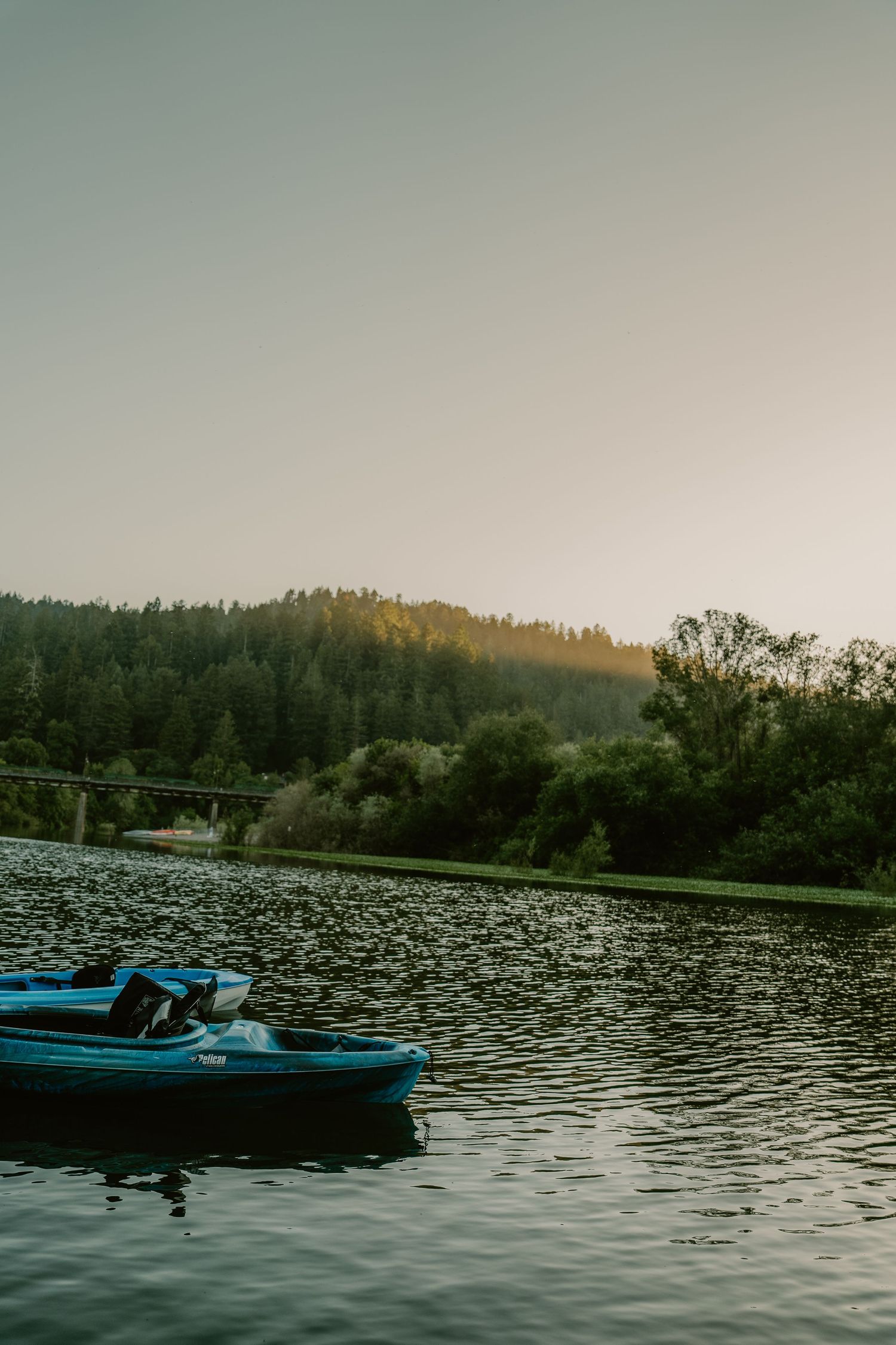 A blue kayak floating on a calm lake at sunset with forested hills and a bridge in the background.