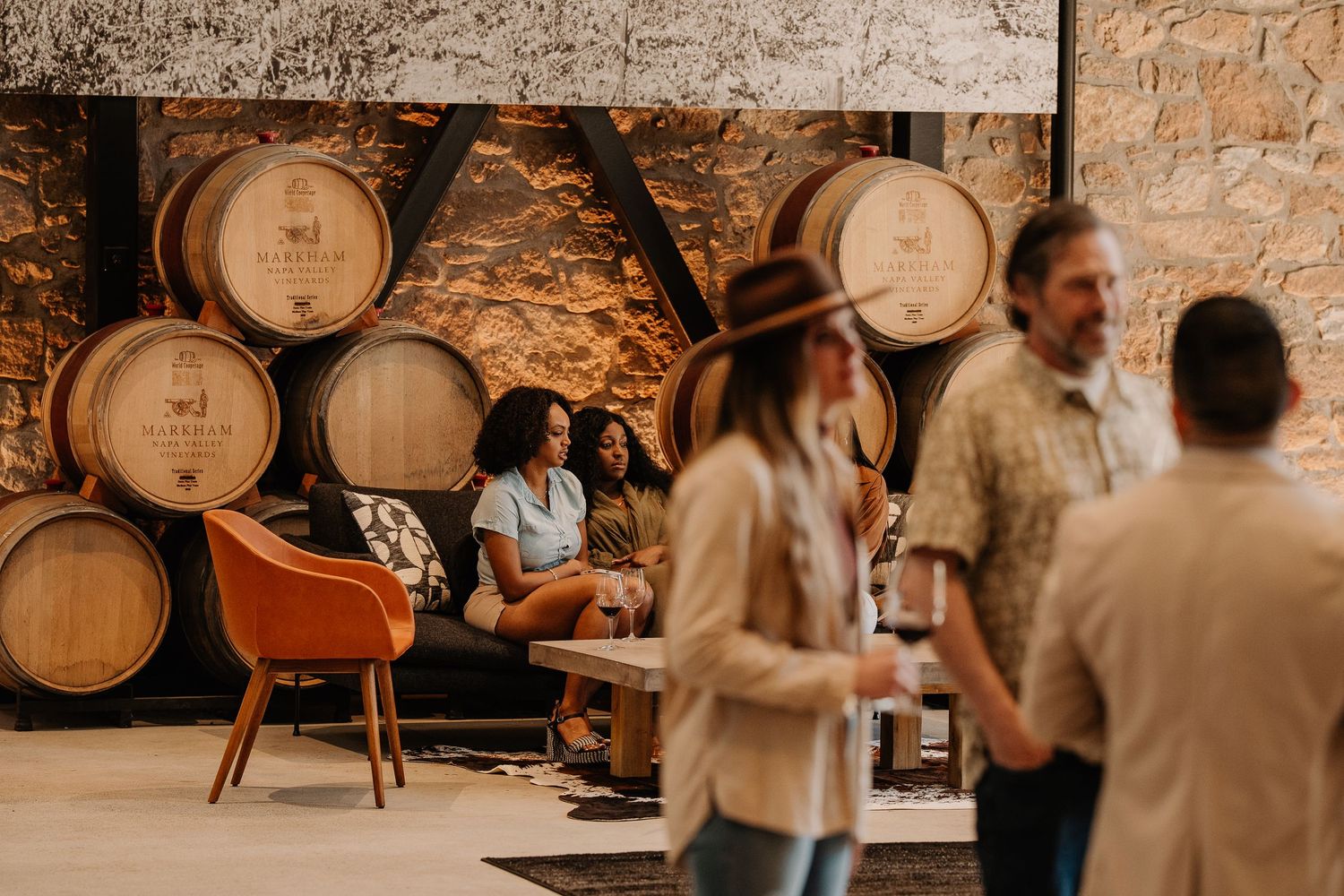 People socializing at a rustic winery tasting room with oak barrels and stone walls in the background.