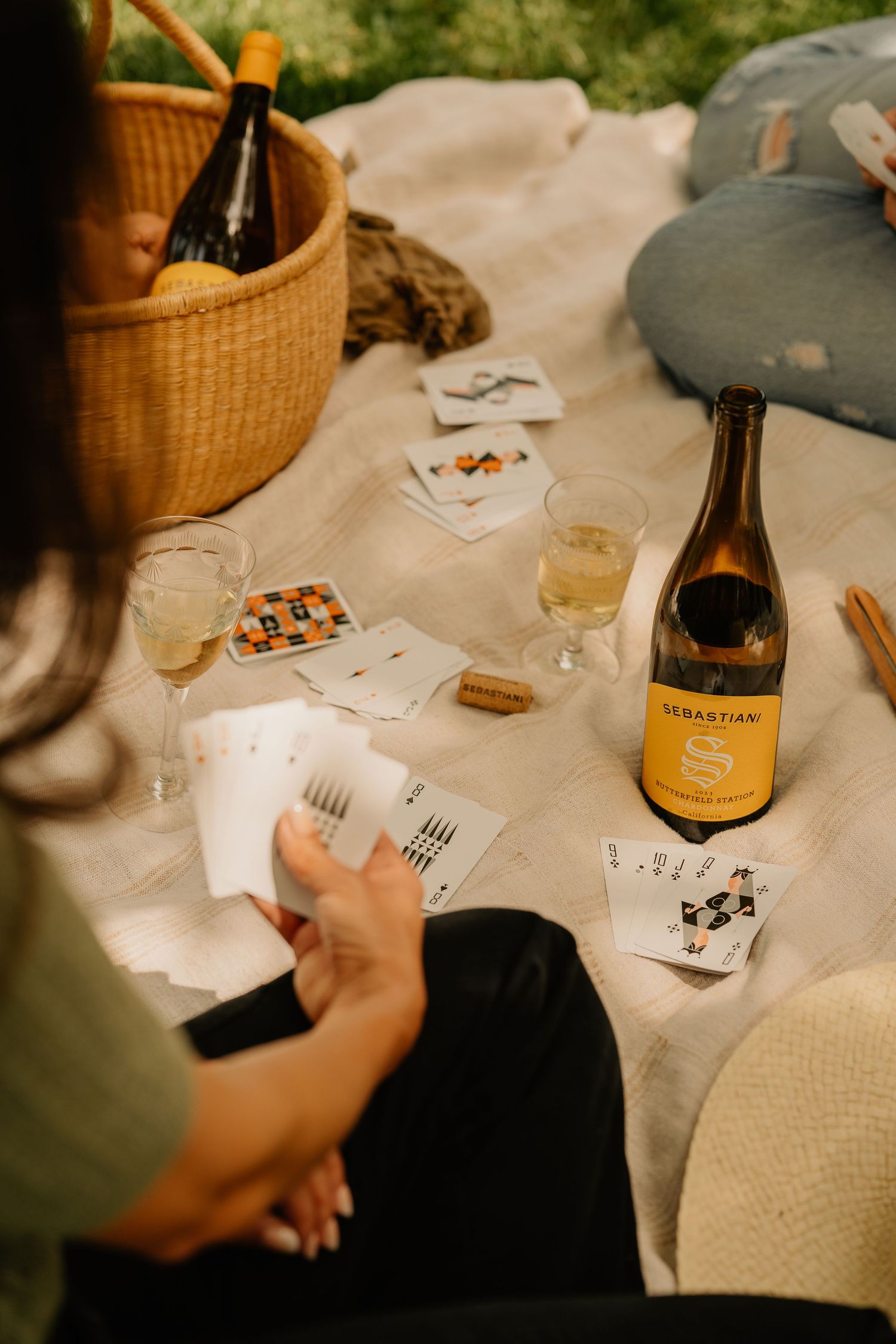 Outdoor picnic scene with Sebastiani Chardonnay, playing cards, wine glasses and wicker basket on blanket.