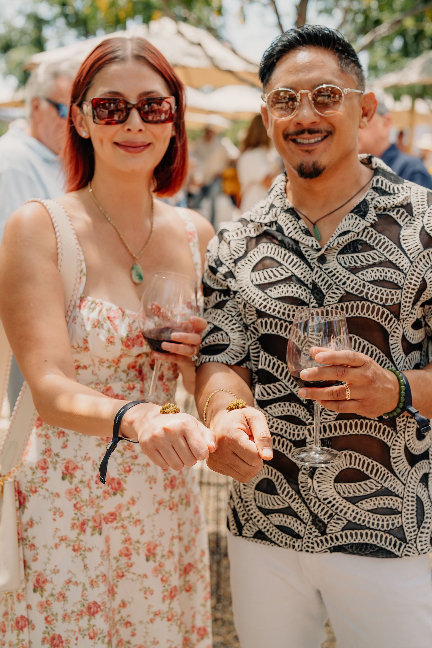 Couple holding hands and smiling at an outdoor wine tasting event, woman in floral dress with sunglasses, man in patterned shirt.