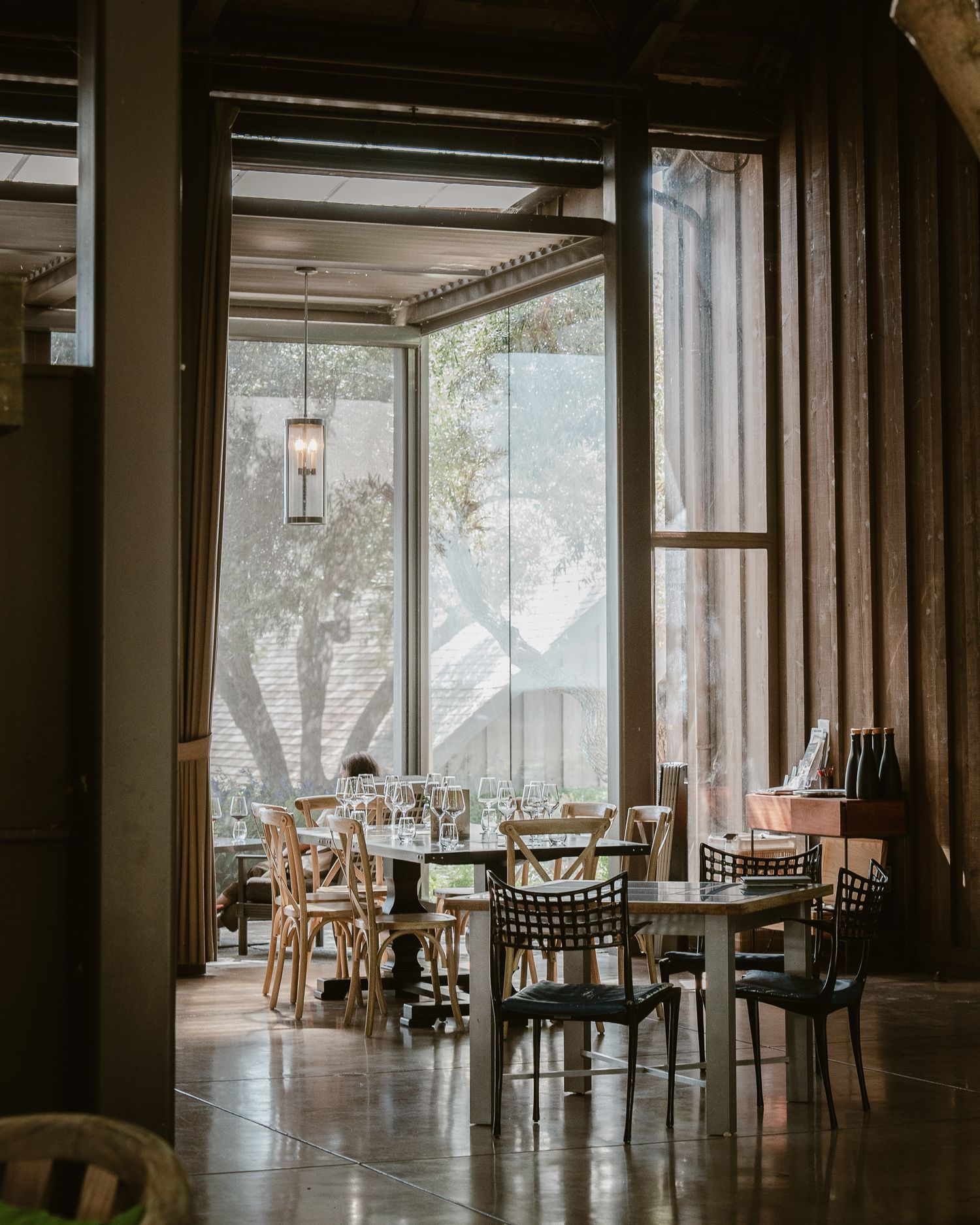 Interior of empty rustic restaurant with wood tables and chairs near large windows letting in natural light.