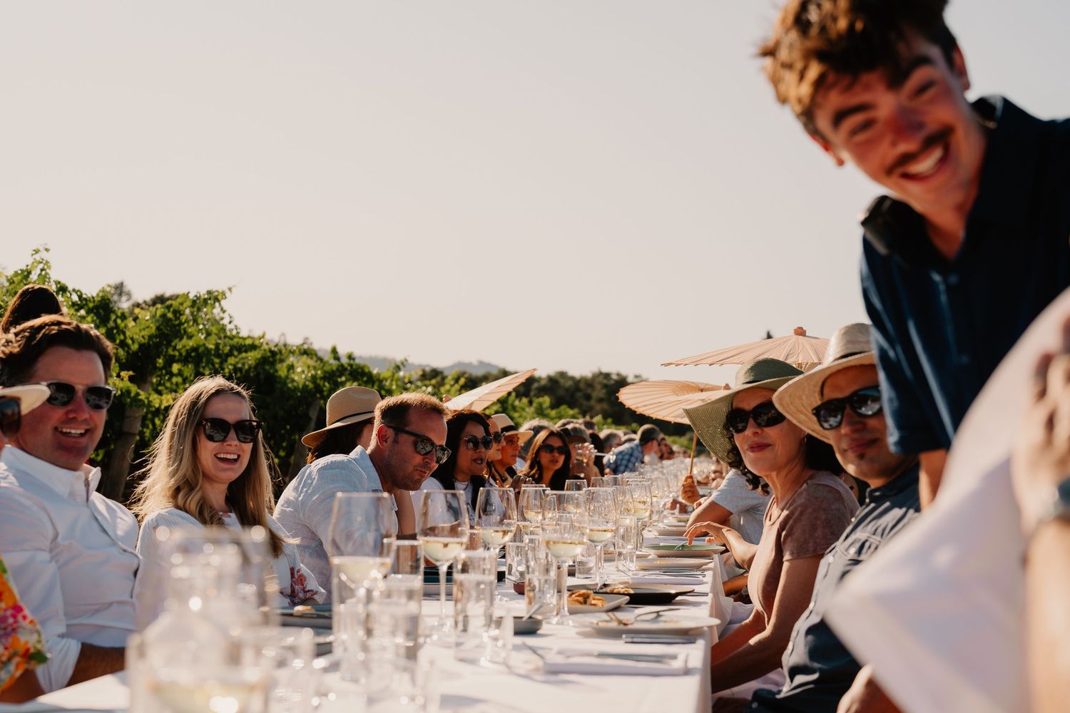 Guests seated at a long outdoor dining table with wine glasses during a sunny afternoon celebration.