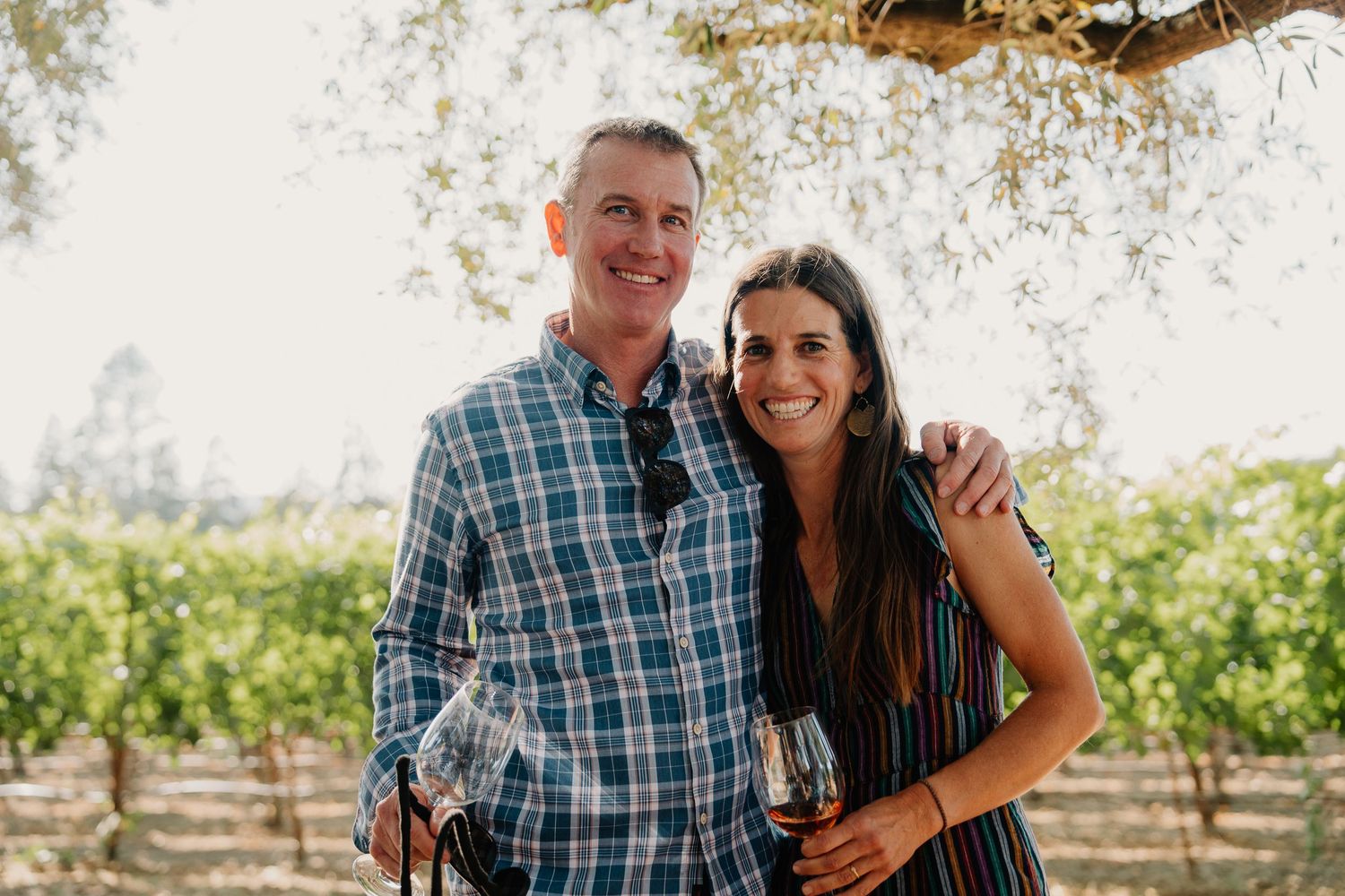 Two people smile together while wine tasting at a sunny vineyard with rows of grapevines in the background.