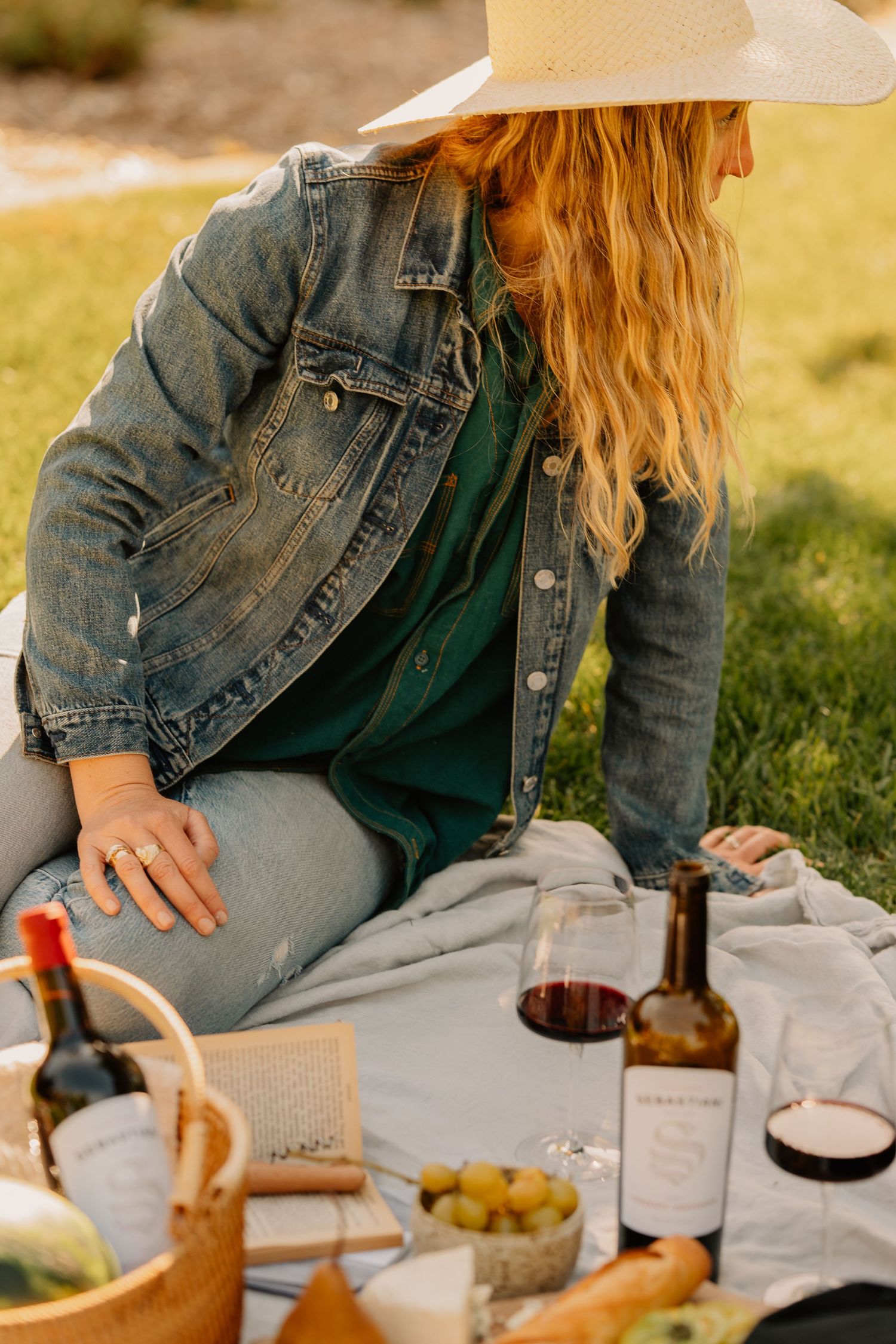 Woman in denim jacket and white hat sitting on a picnic blanket with red wine and cheese.
