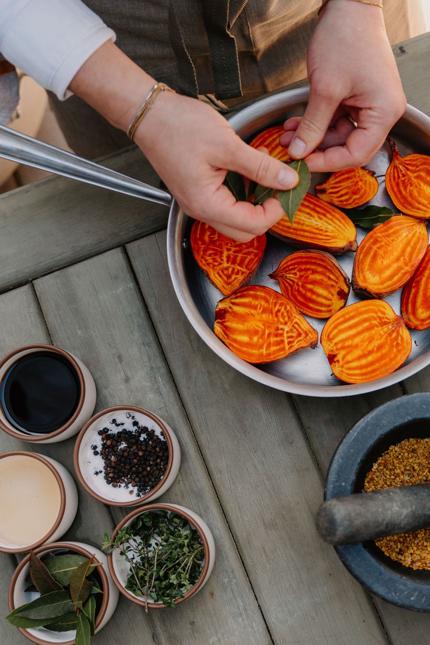 Hands placing bay leaves on sliced golden beets in a pan, surrounded by spices, herbs, and a mortar on a wooden table.