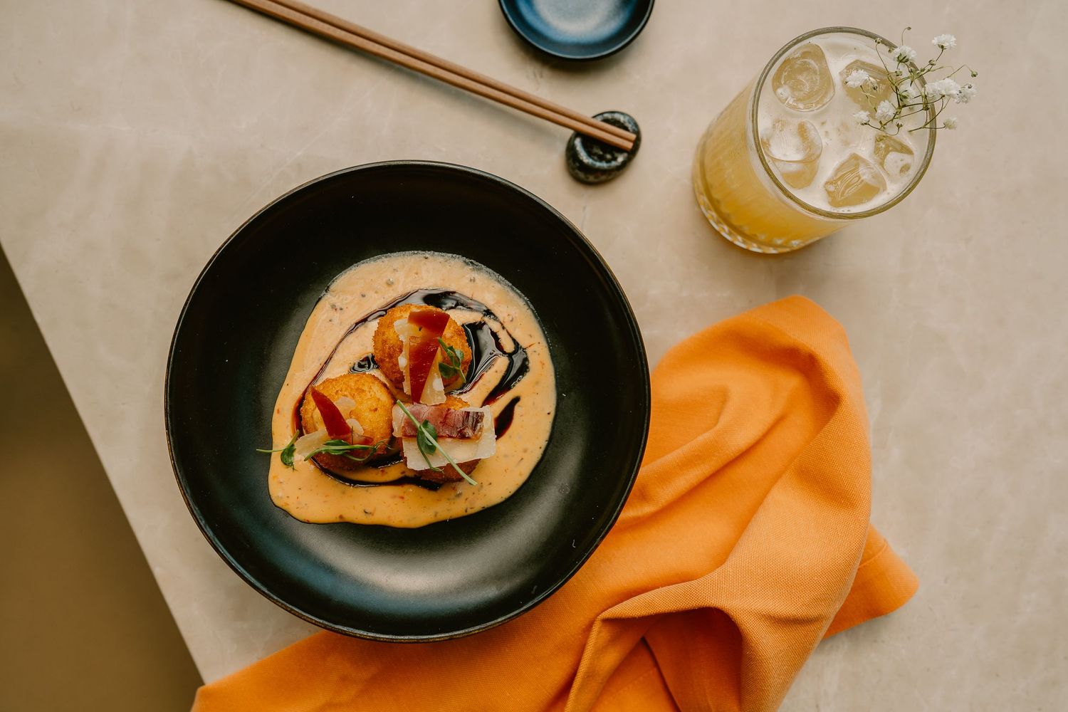Overhead view of a gourmet dish in a black bowl with colorful vegetables, beside an iced drink and orange napkin.