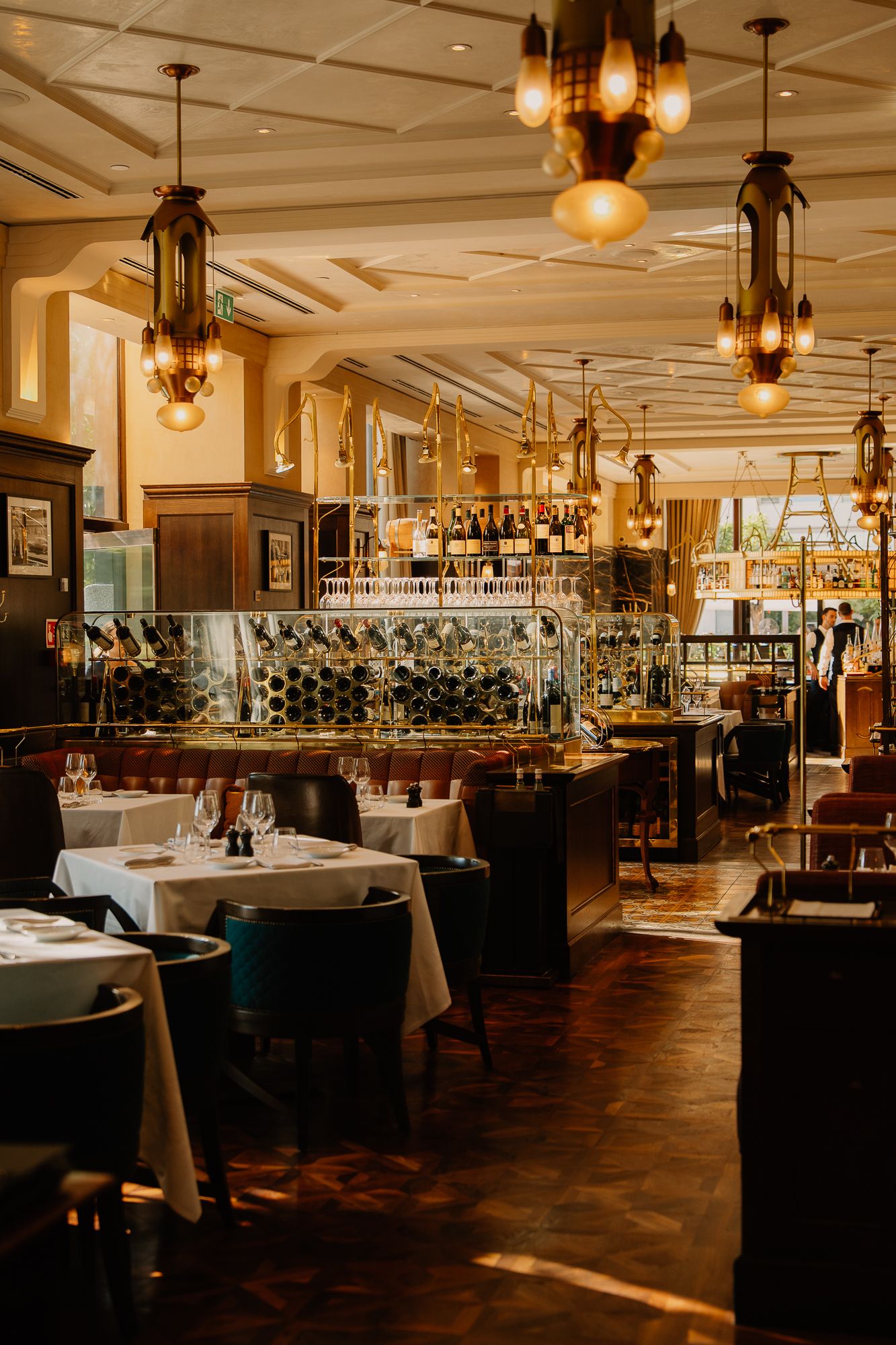 Elegant restaurant interior with white tablecloths, ornate hanging lanterns, and a wine display wall in warm golden lighting.