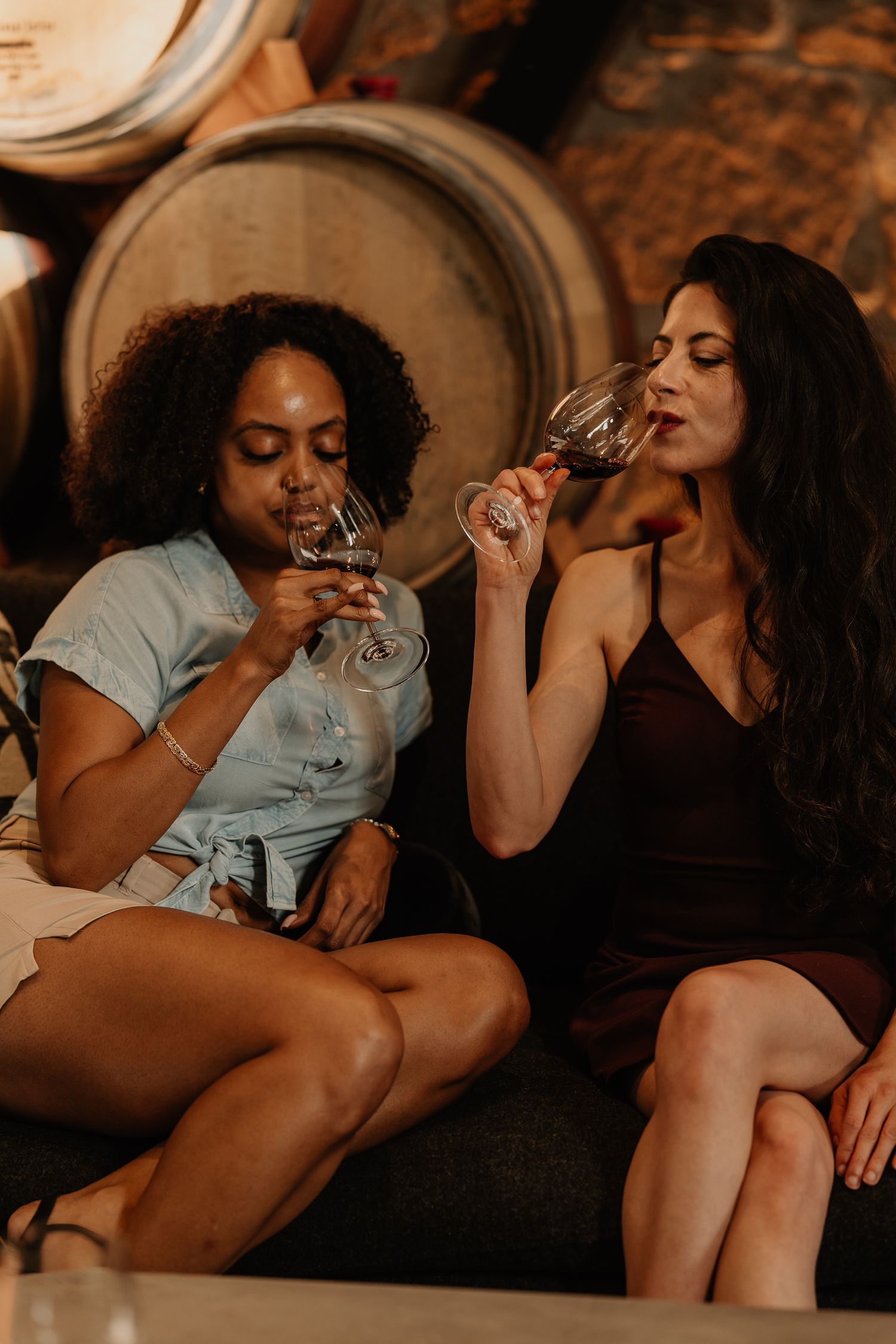 Two women sipping red wine from glasses while seated together in a dimly lit barrel room winery.