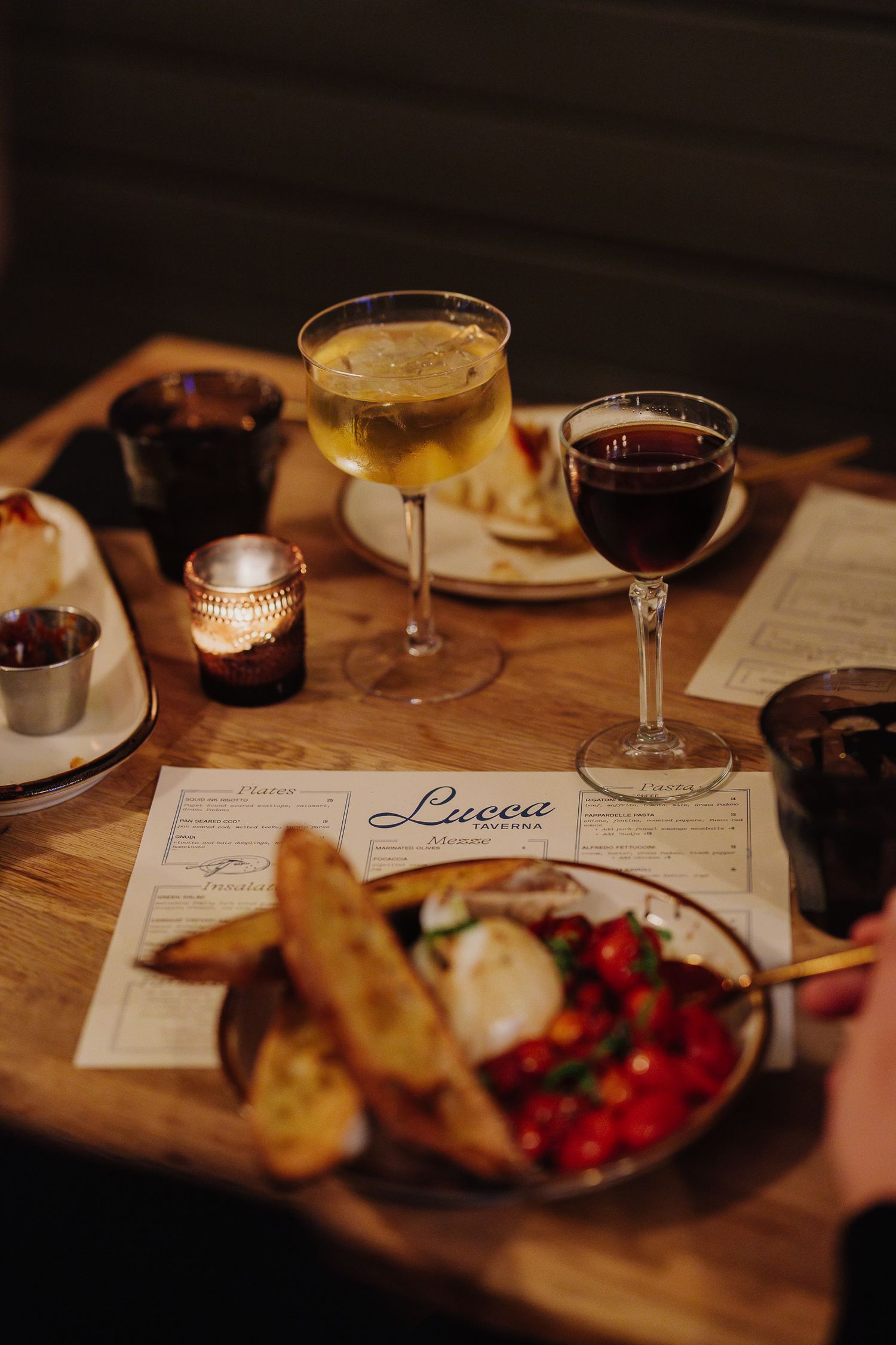 A cozy restaurant table with wine glasses, cocktails, and appetizer plates featuring burrata and bread on a wooden surface.
