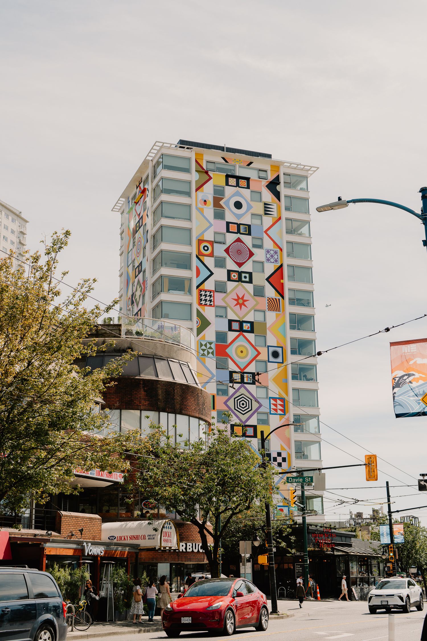 A colorful geometric mural decorates the side of a tall apartment building in an urban street setting.