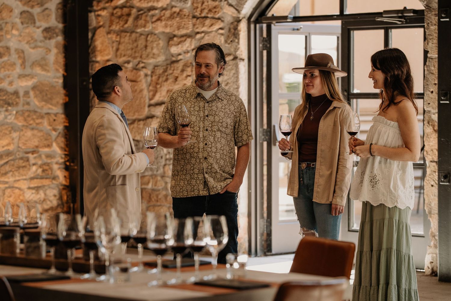 Four guests enjoy a wine tasting in a rustic stone-walled winery, chatting and holding red wine glasses.