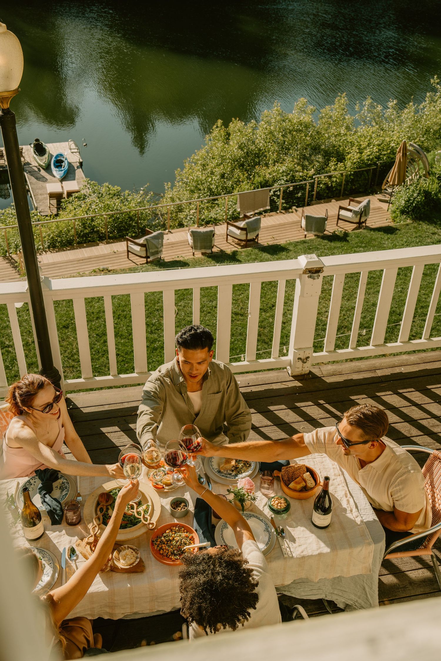 Friends dining al fresco on a sunny deck overlooking a serene river, sharing food and wine together.