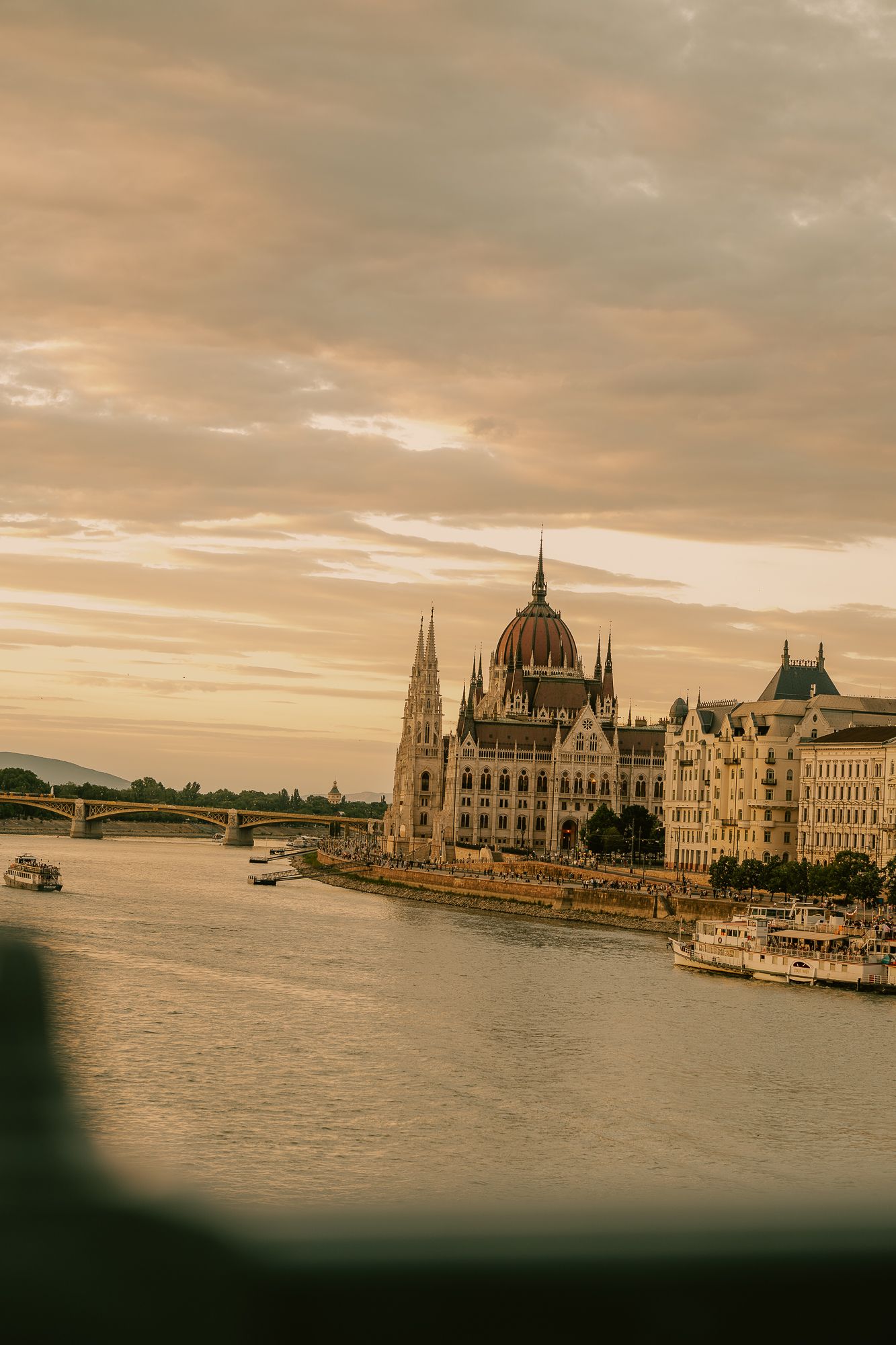 Golden sunset over the Hungarian Parliament Building along the Danube River in Budapest, with boats and a bridge visible.