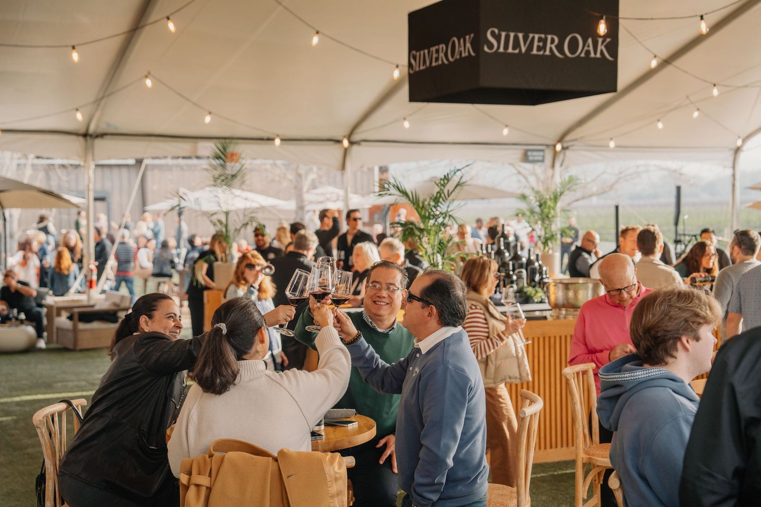 Crowd of guests toasting and enjoying wine at a busy Silver Oak winery event under a branded tent.