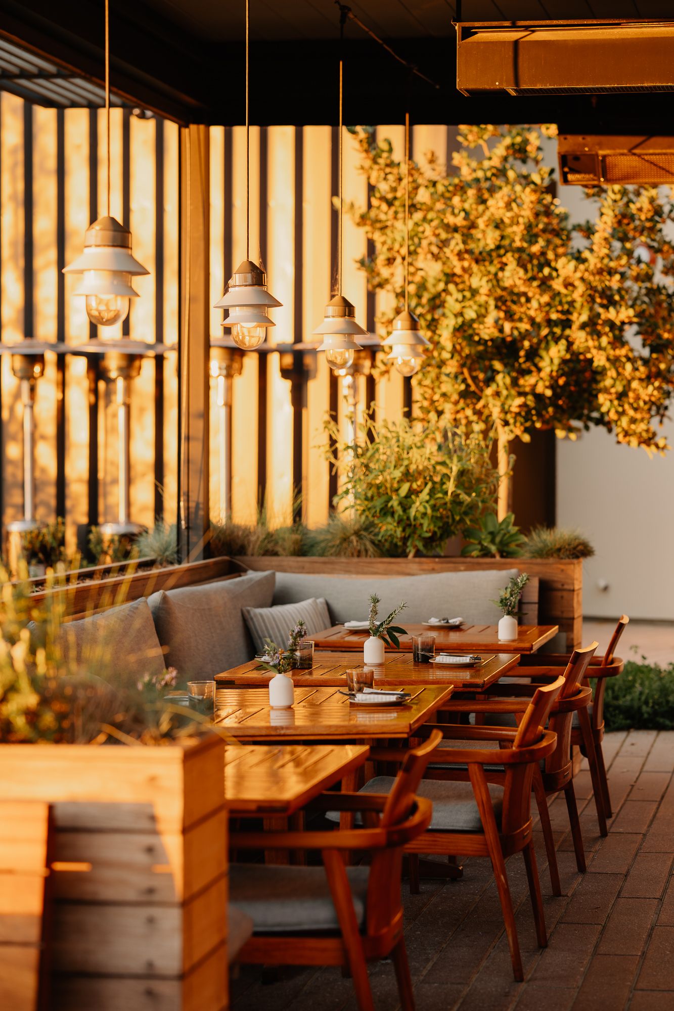 Warm outdoor restaurant patio with wooden tables, pendant lights, and lush greenery in golden evening light.
