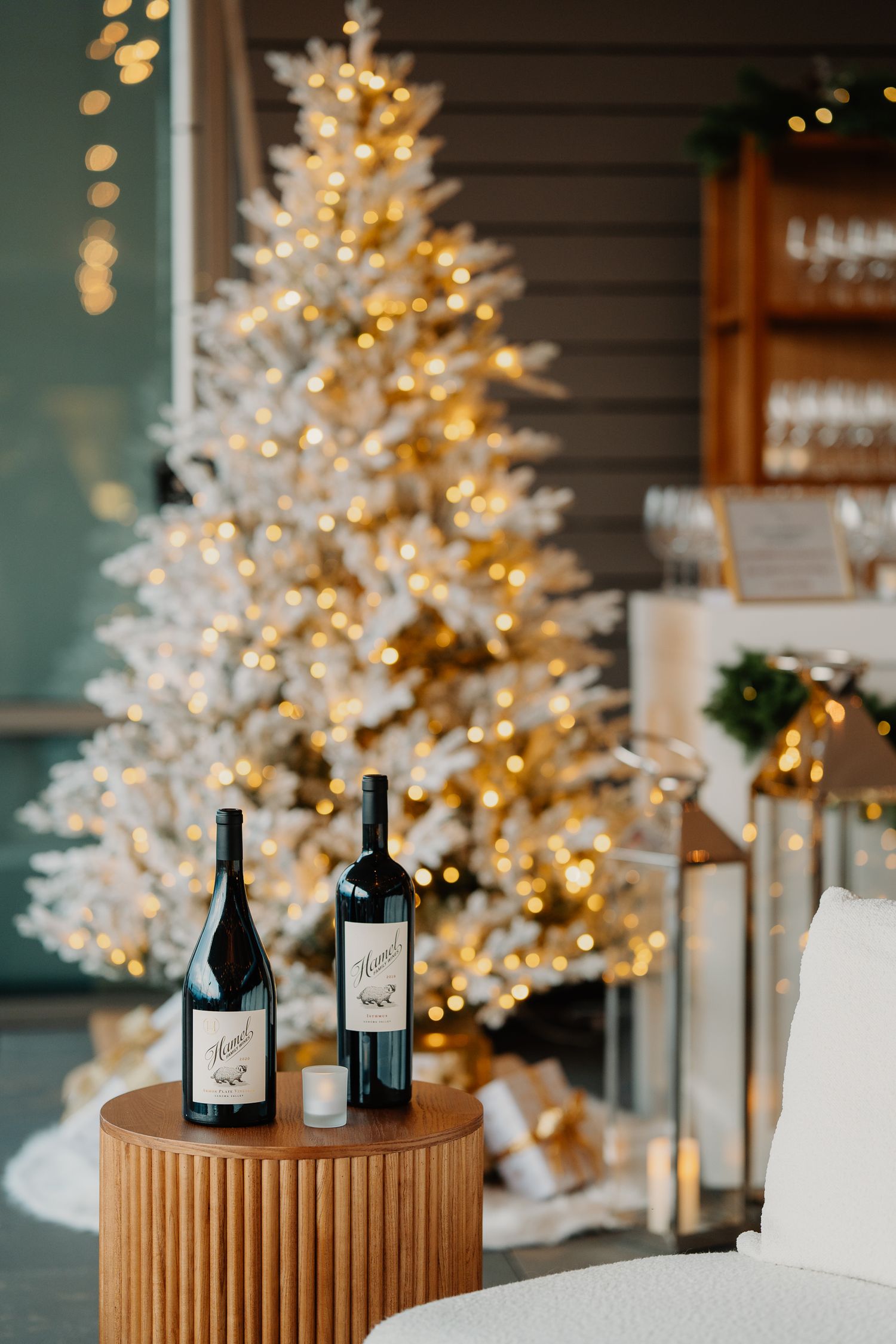 Wine bottles displayed on a round wooden pedestal with a white illuminated Christmas tree glowing in the background.