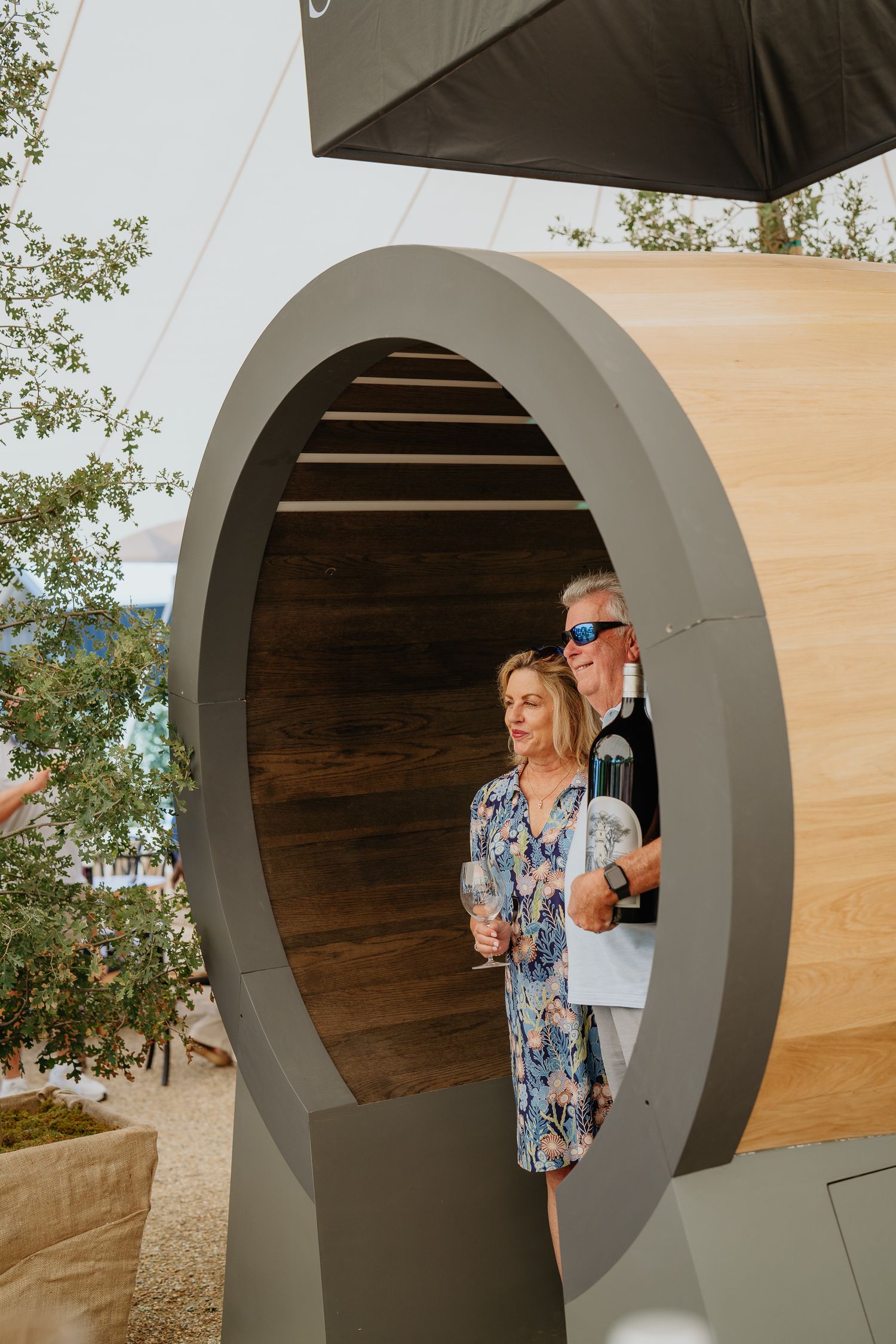 Couple holding wine glasses inside a modern oval-shaped wine barrel installation at an outdoor winery event.