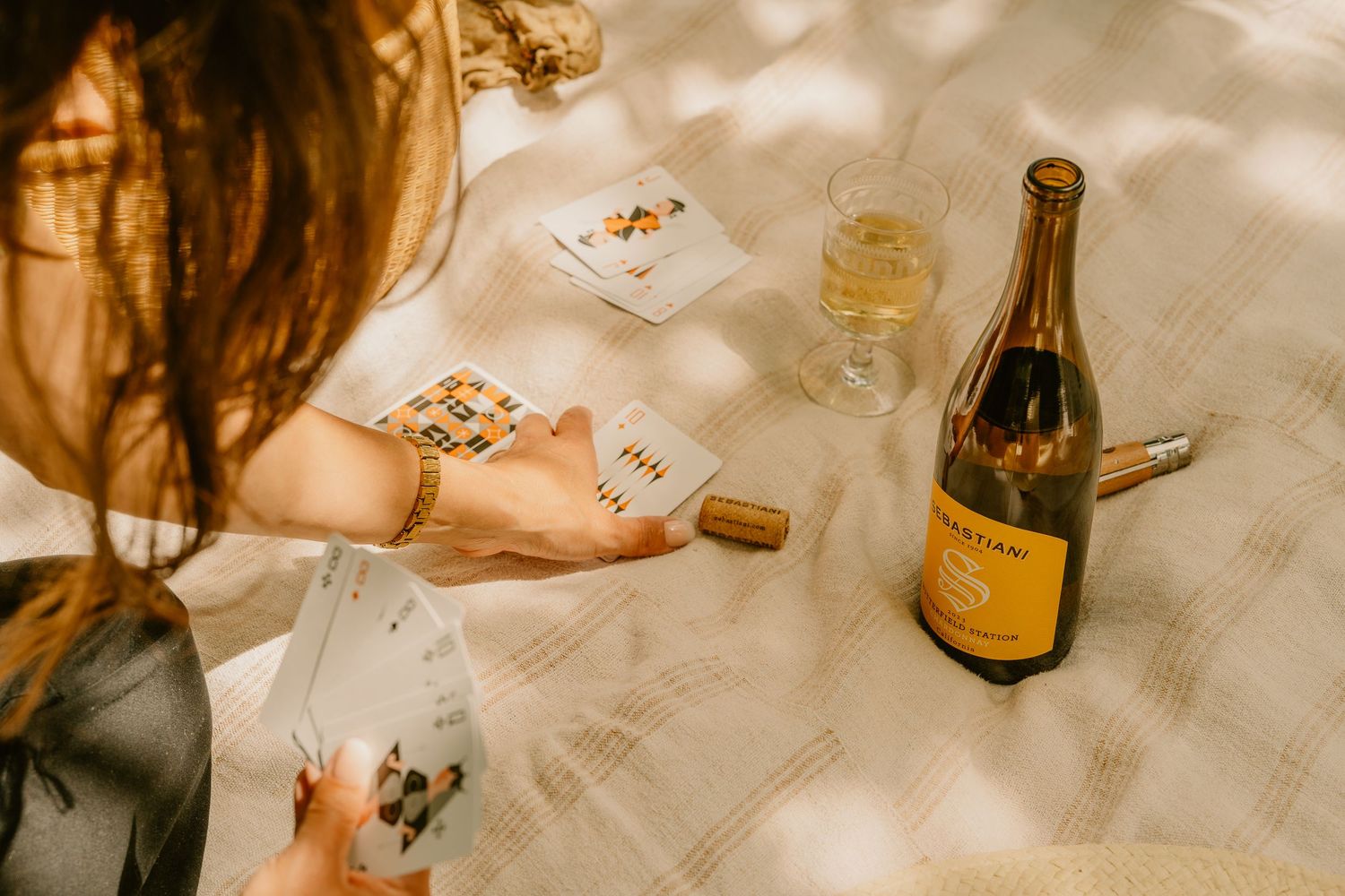 Woman playing cards at a picnic with a bottle of Sebastiani sparkling wine and a glass on a linen blanket.