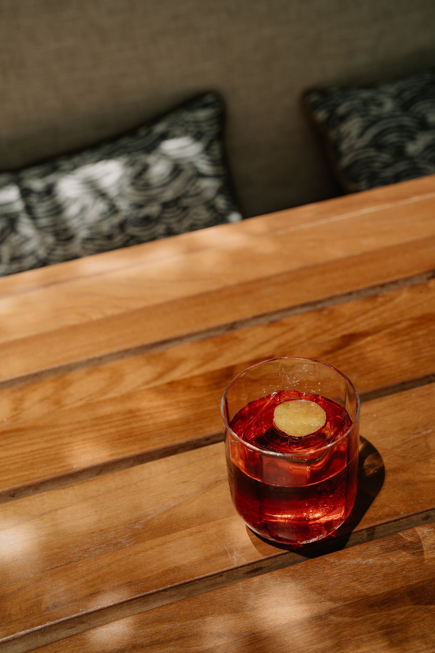 A glass of red cocktail with a lemon garnish sits on a rustic wooden table in dramatic lighting.