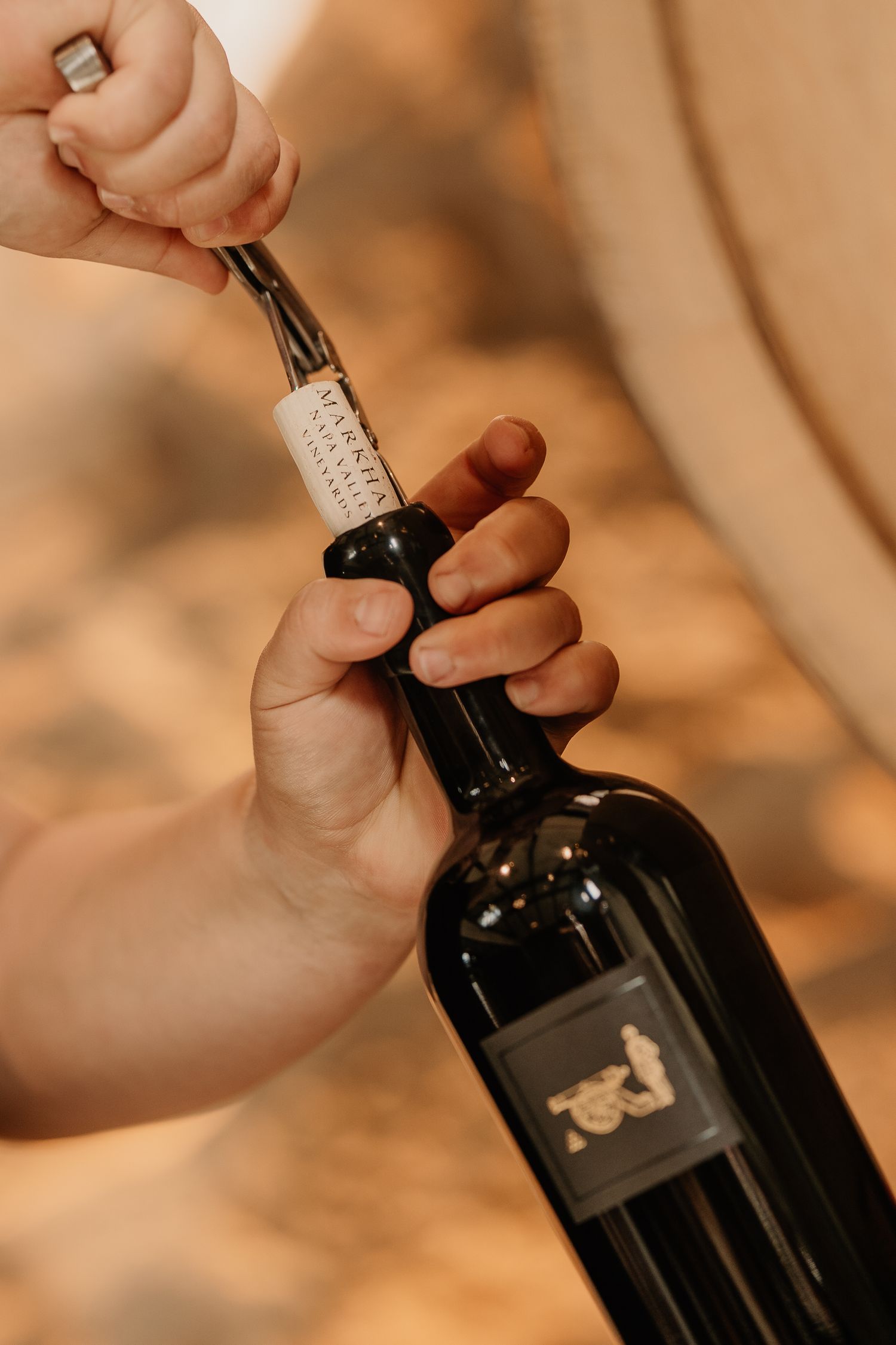Hands using a corkscrew to open a dark wine bottle with a black label in a warm cellar setting.