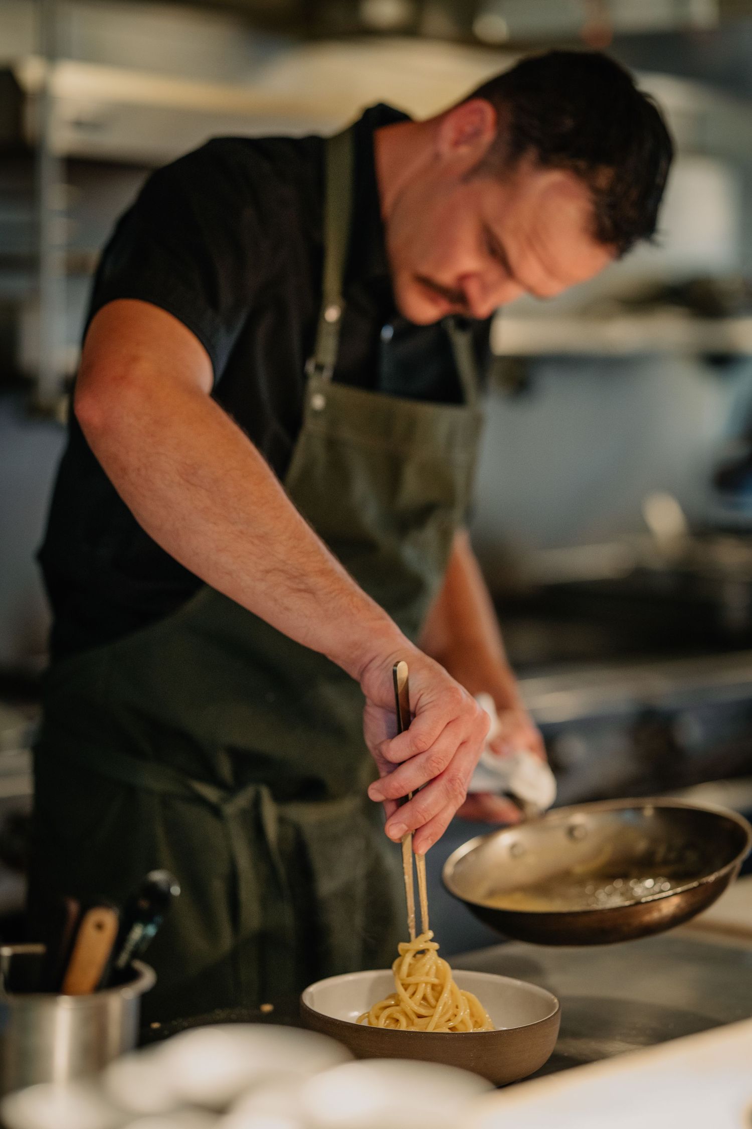 Chef in green apron plating fresh spaghetti pasta into a bowl using tongs in a professional restaurant kitchen.