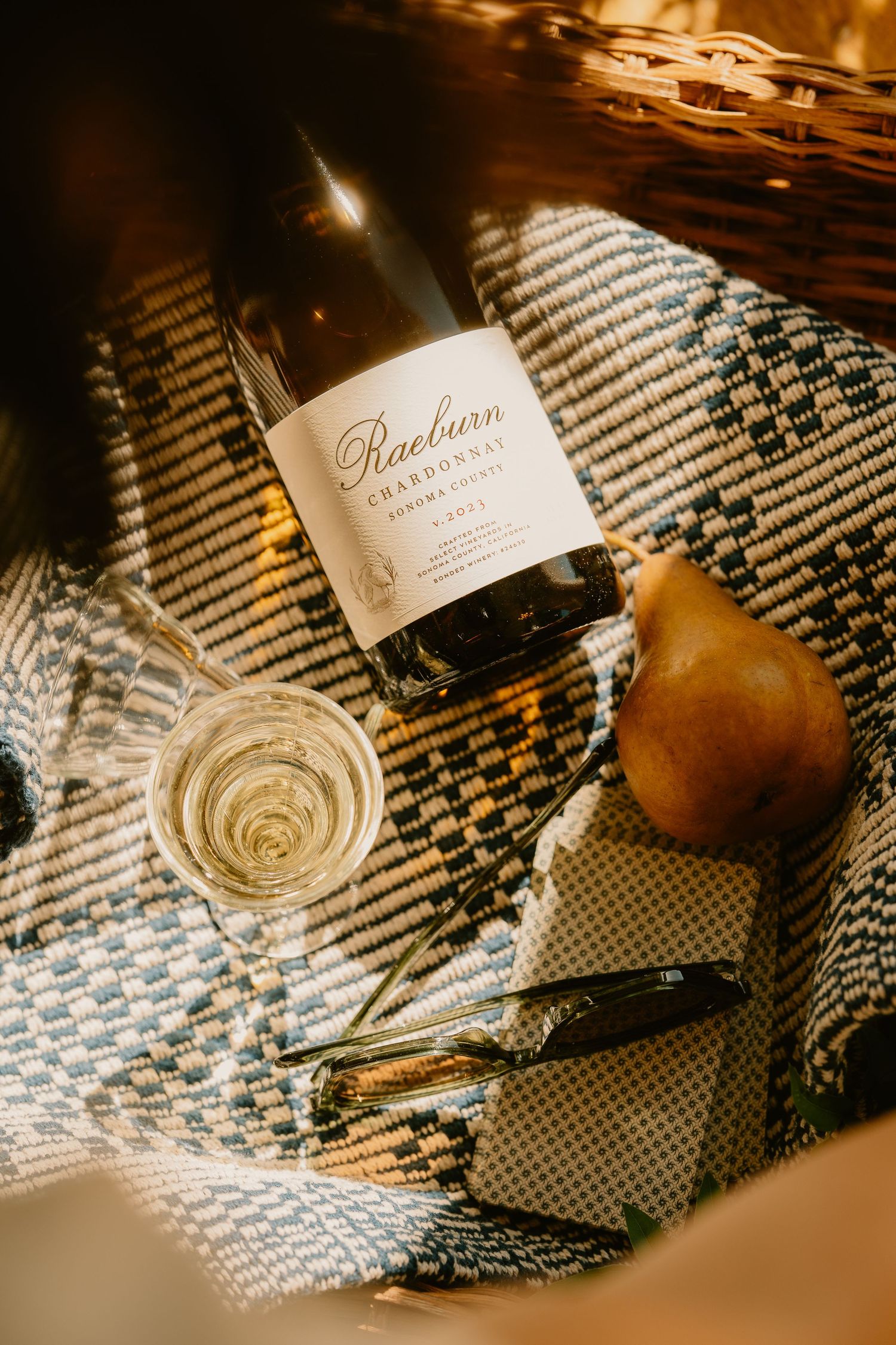 Raeburn Chardonnay bottle with a wine glass, pear, and sunglasses on a woven blanket in warm light.