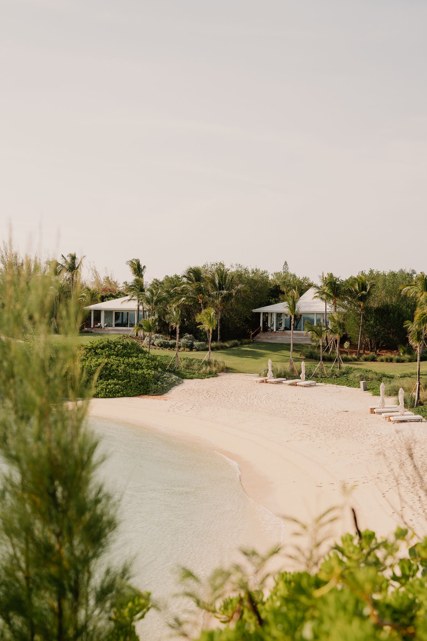 A serene tropical beachfront view with white sand, turquoise water, palm trees and coastal cottages along the shore.